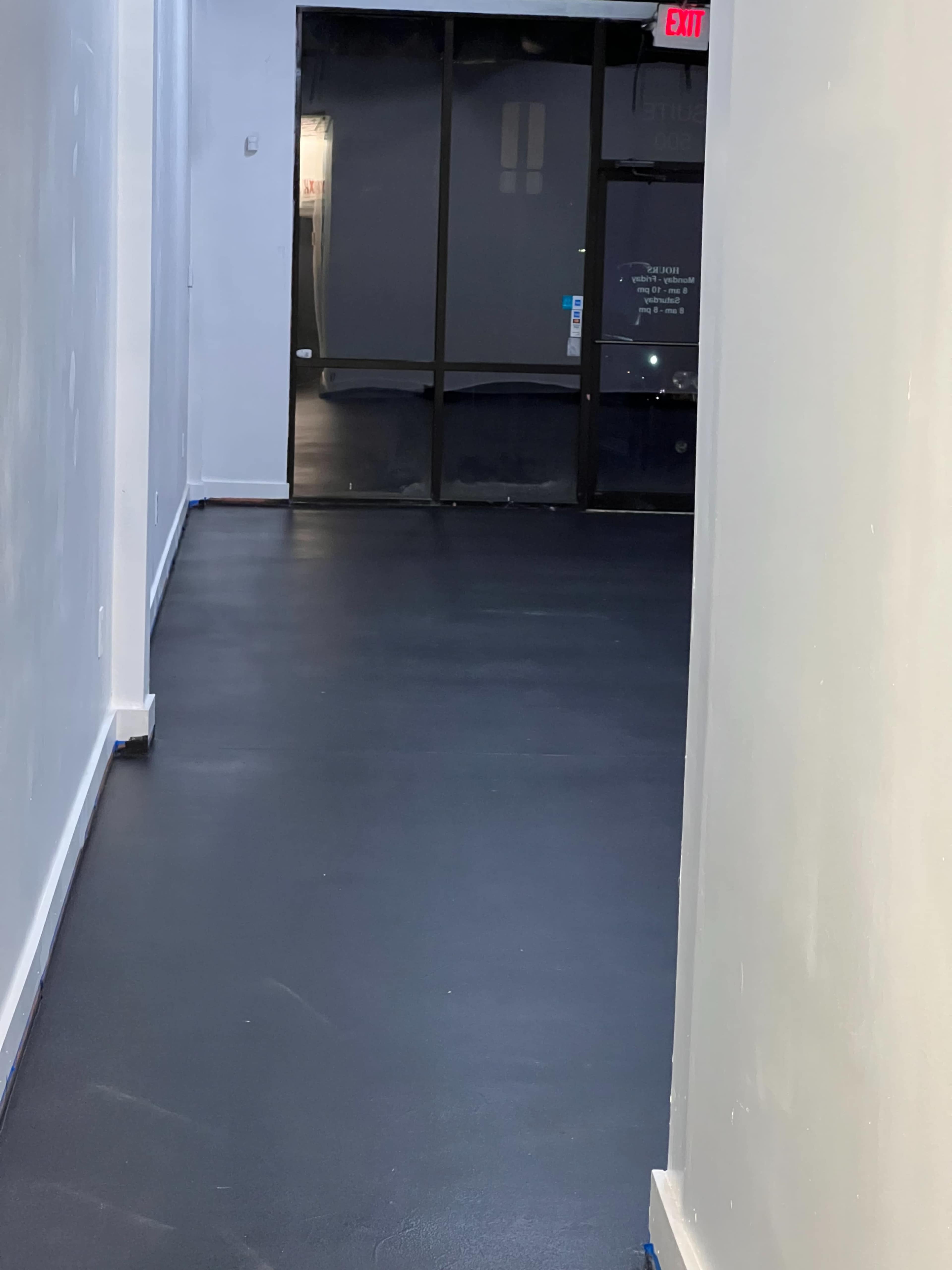 The image shows a narrow corridor with black flooring leading to a glass door at the far end.