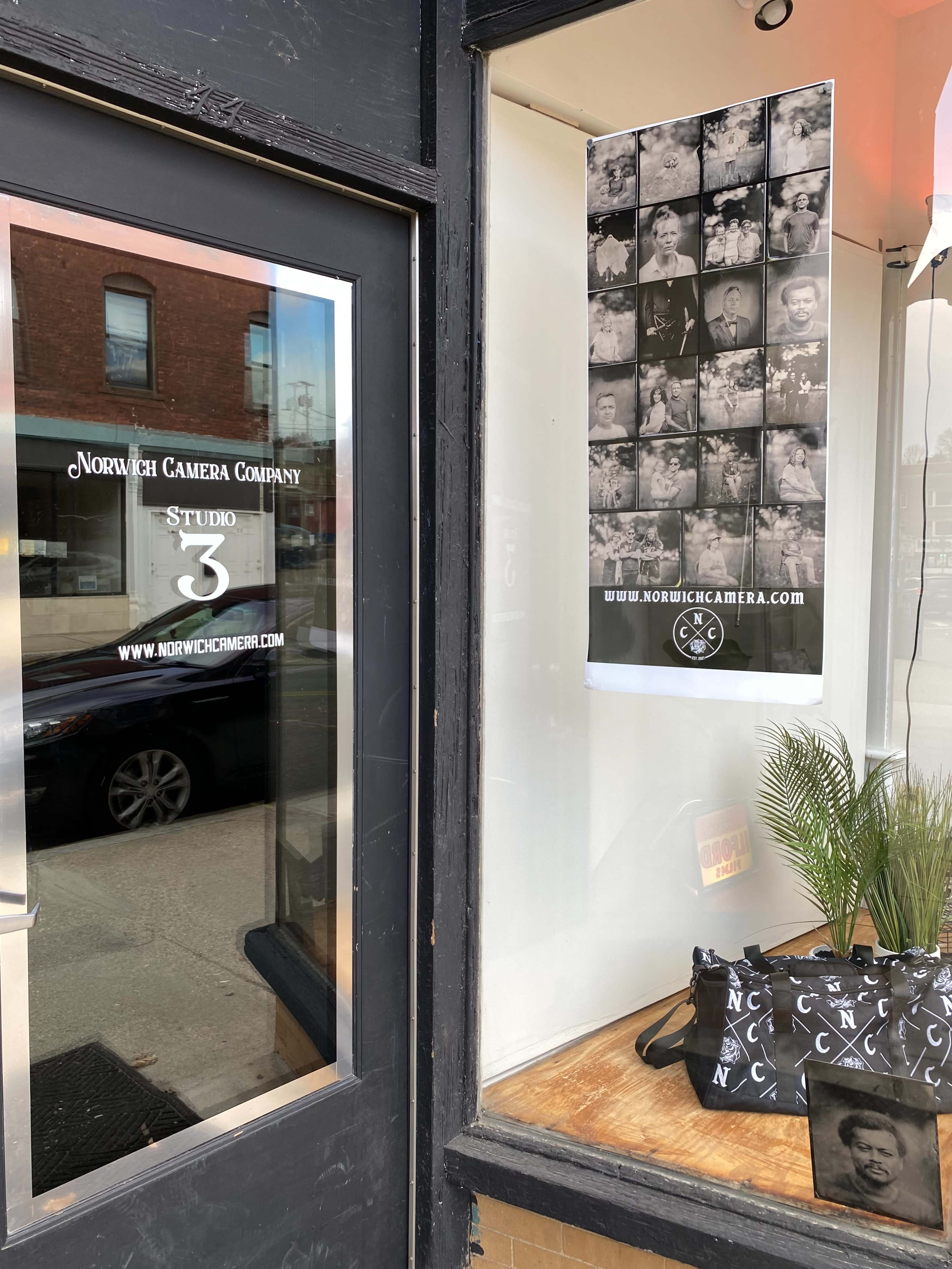The image shows the exterior of the Norwich Camera Company, featuring a window display with a poster and a decorative plant.