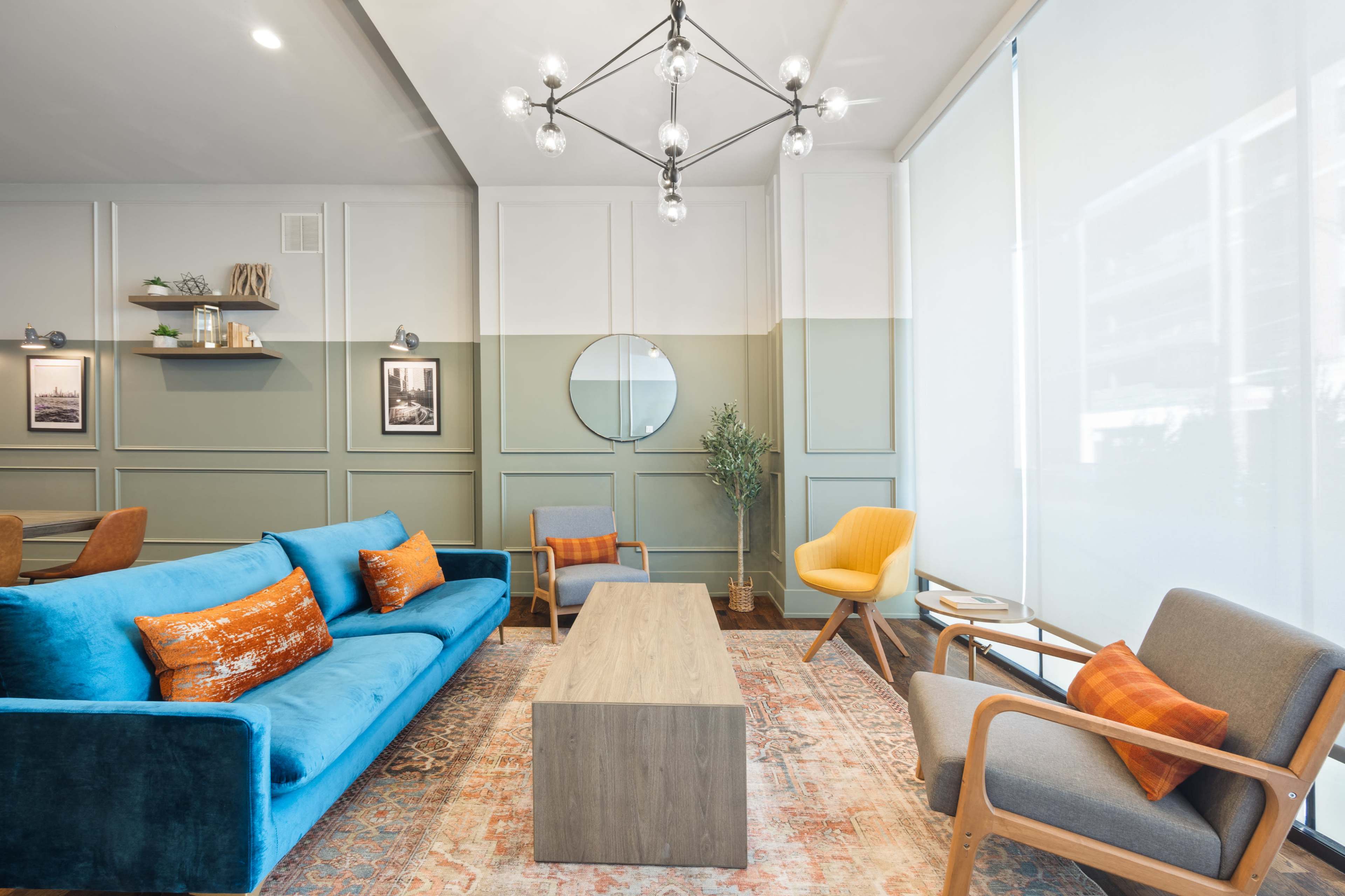 A modern lounge area featuring a blue velvet sofa, a wooden coffee table, and a mix of armchairs, with decorative wall frames and a round mirror.