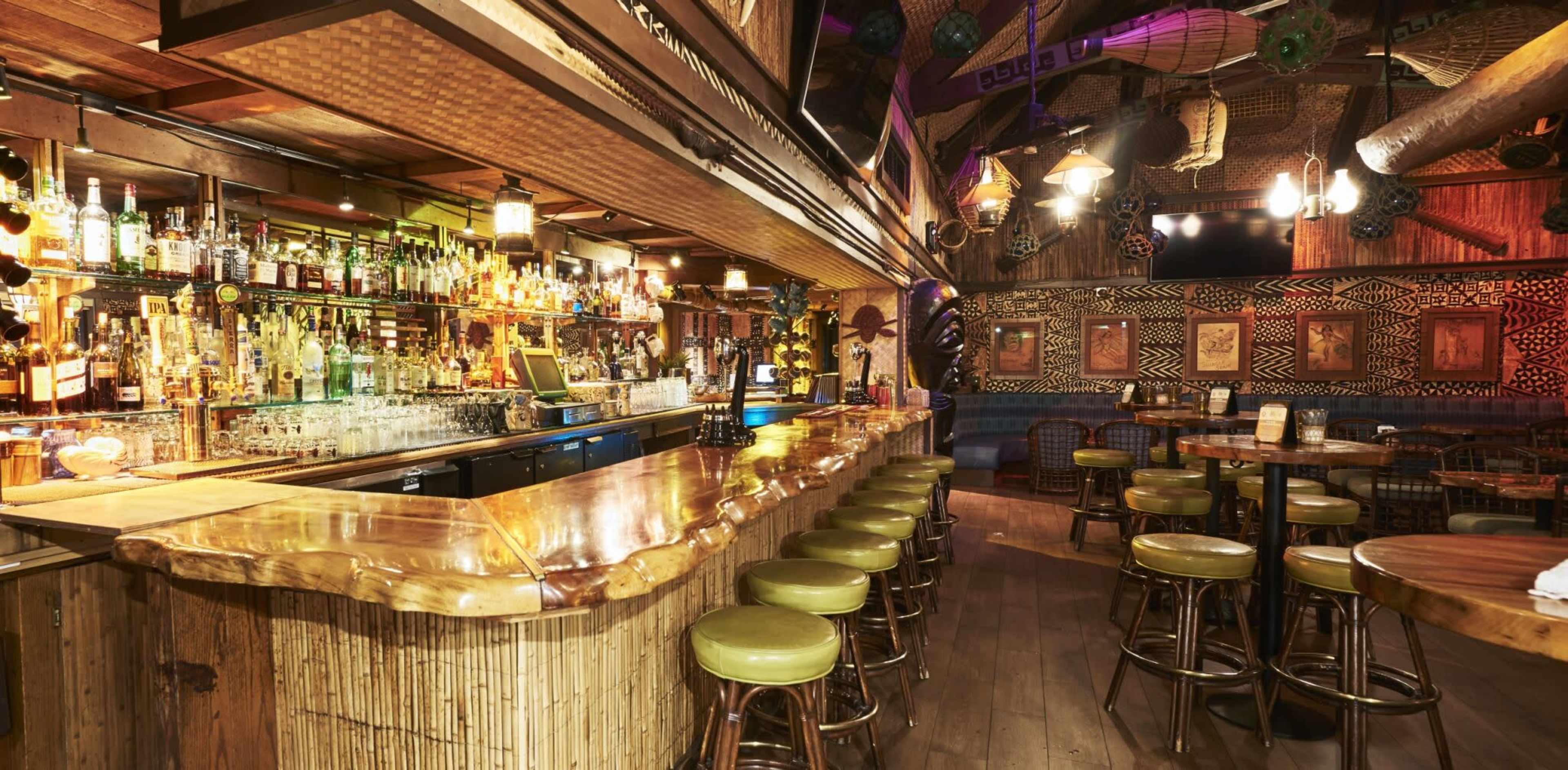 The image shows a brightly lit bar with a bamboo counter, numerous bottles on shelves, and circular green stools arranged along the bar.