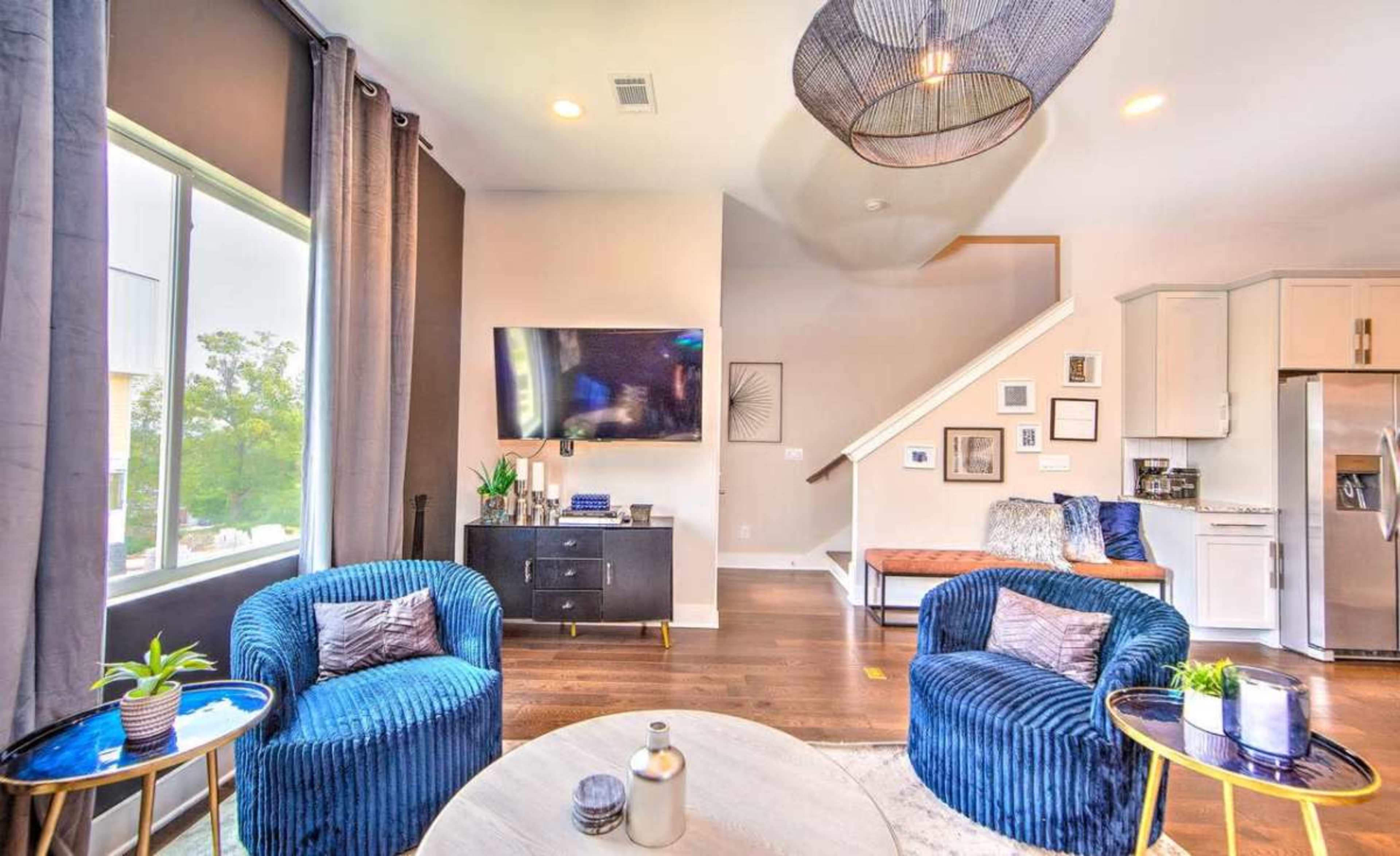 The image features a modern living room with two blue armchairs, a round coffee table, a television mounted on the wall, and a staircase in the background.