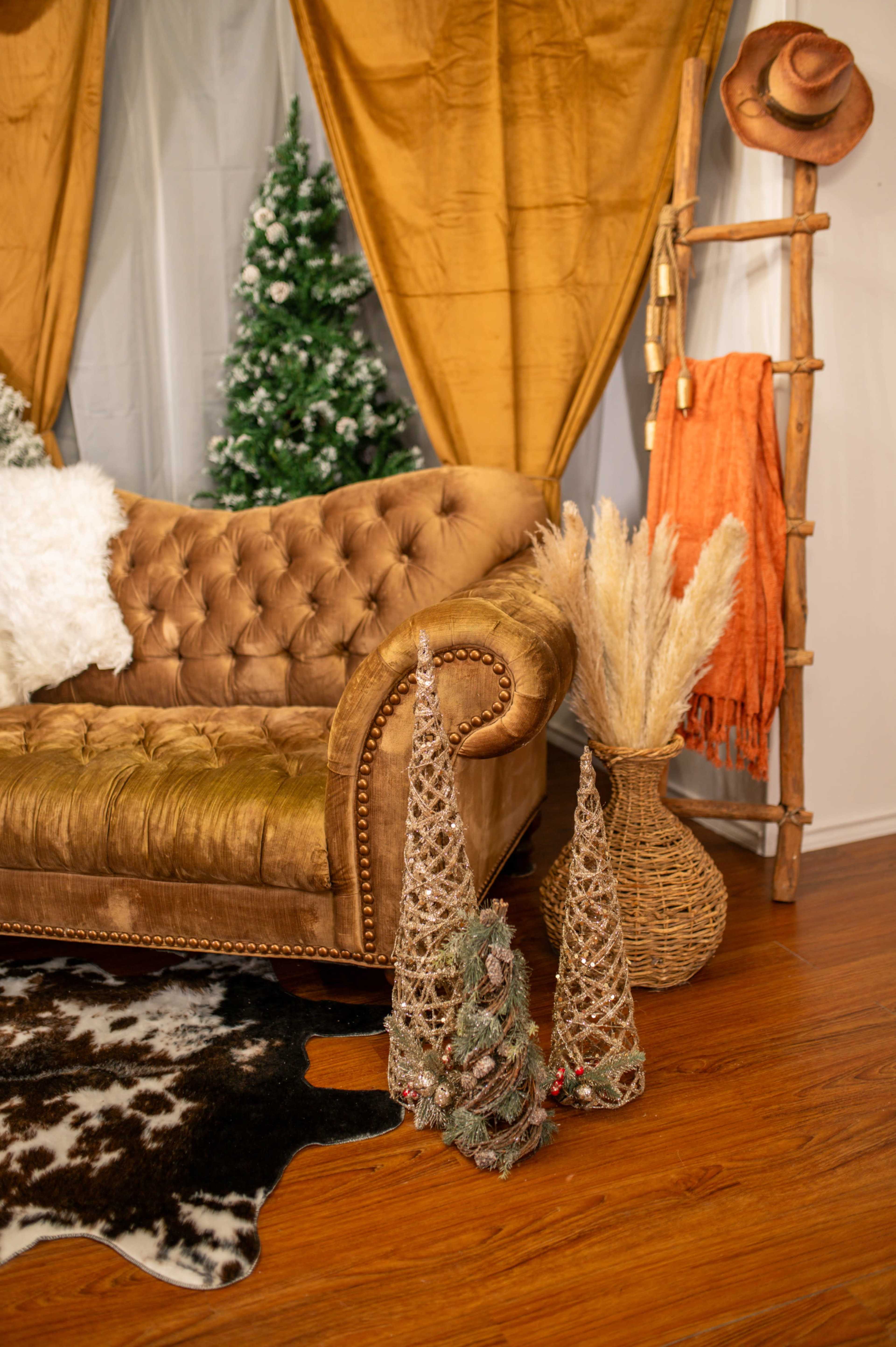 The image features a cozy living space with a brown tufted sofa, decorative trees made of woven material, a wooden rack holding an orange throw, and a faux cowhide rug on the floor.