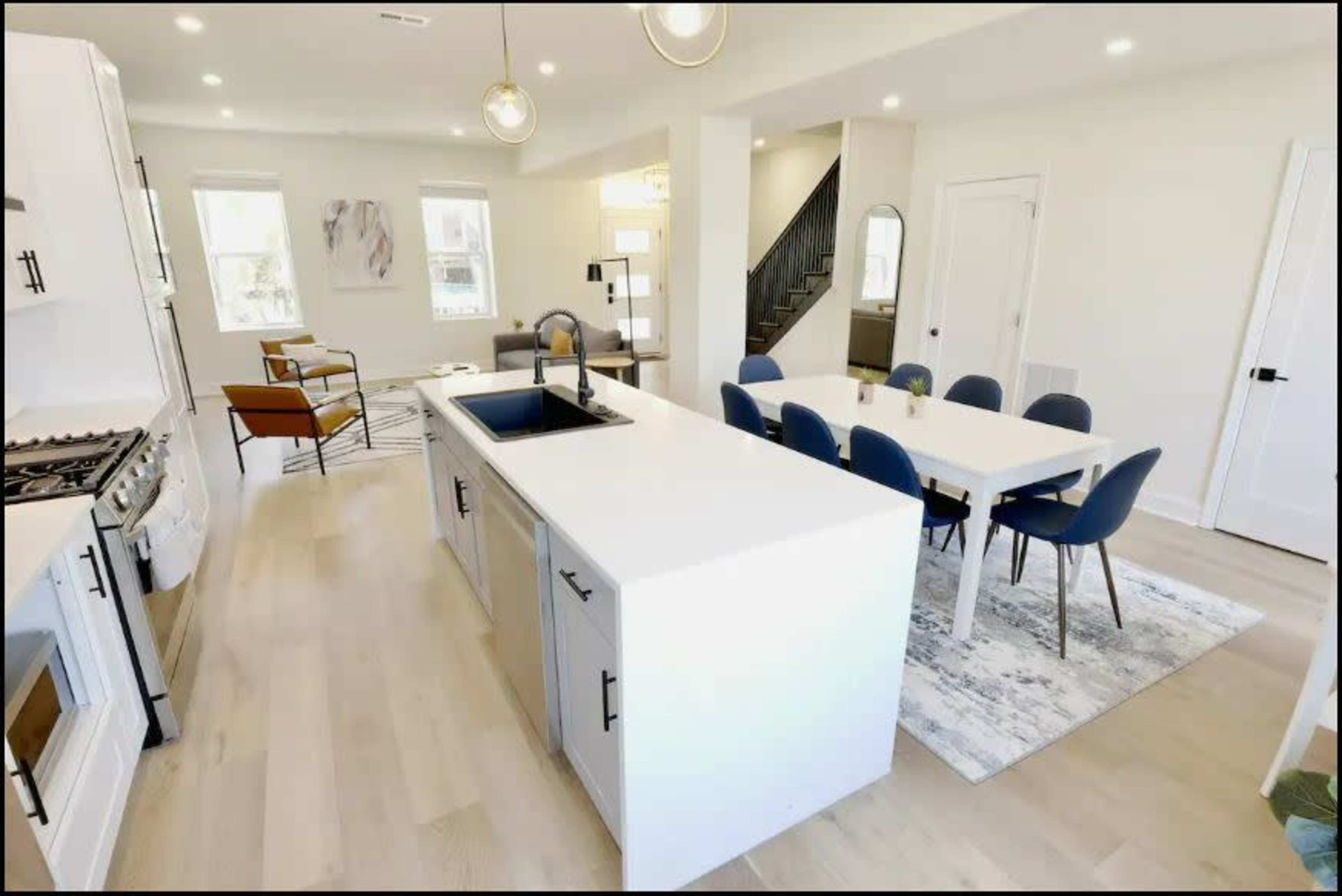 A modern kitchen and dining area with a central island, a dining table with blue chairs, and a view of a staircase in a well-lit space.