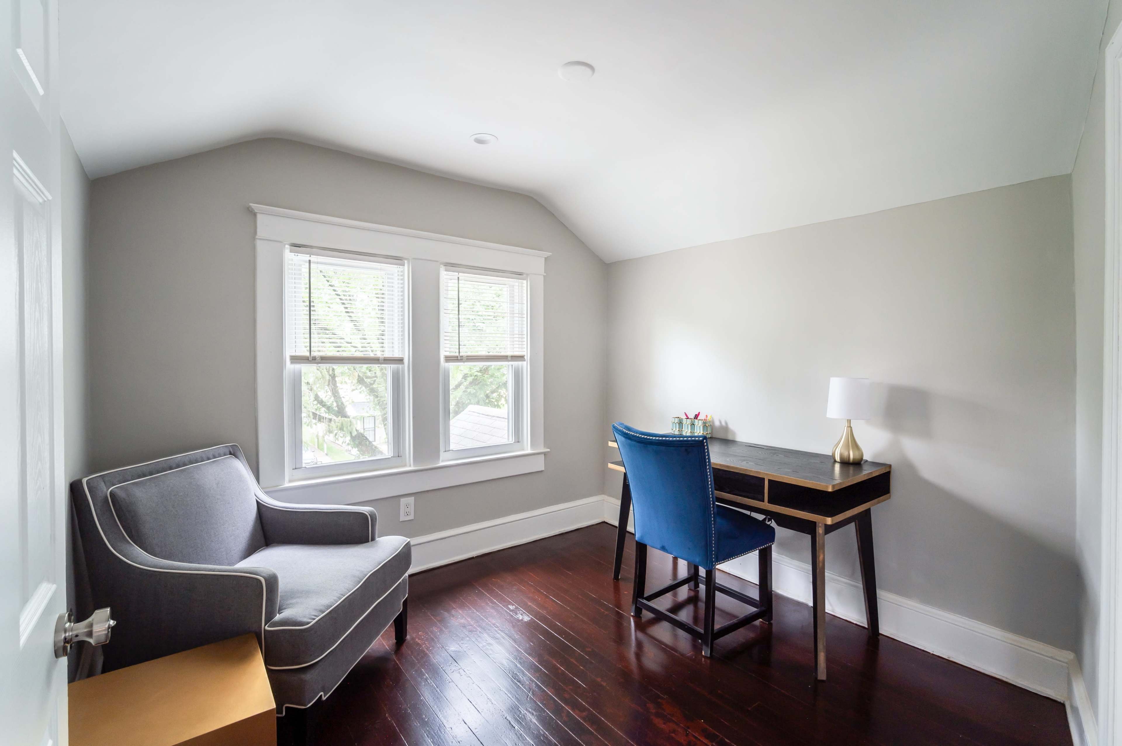 A small, light-filled room with a grey armchair, a blue desk chair, a wooden desk, and large windows.