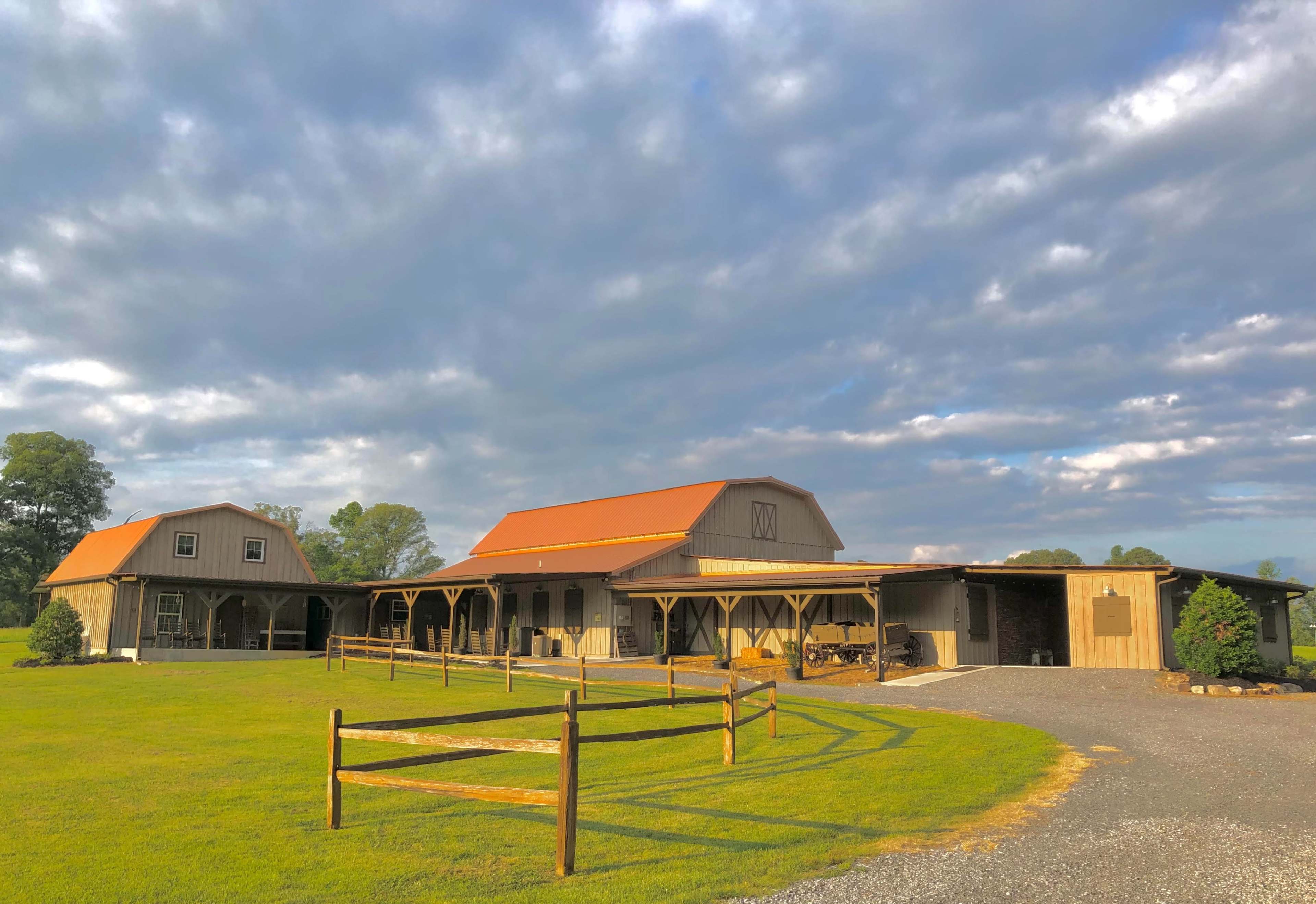 A rural property features a large barn-style building with a reddish-orange roof, set against a backdrop of cloudy skies and green fields.