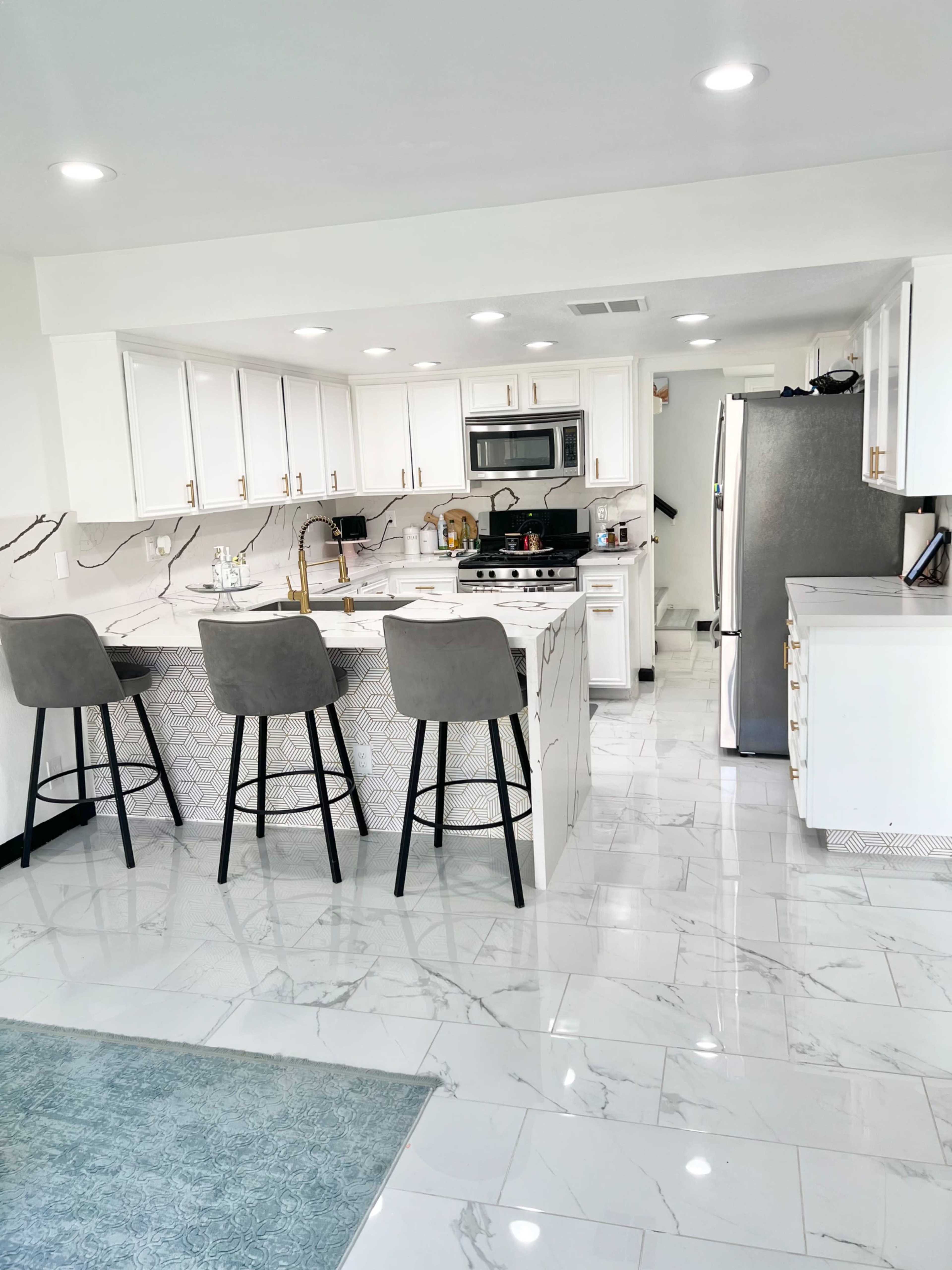 A modern kitchen with white cabinets, gray bar stools, stainless steel appliances, and a marble-patterned backsplash.