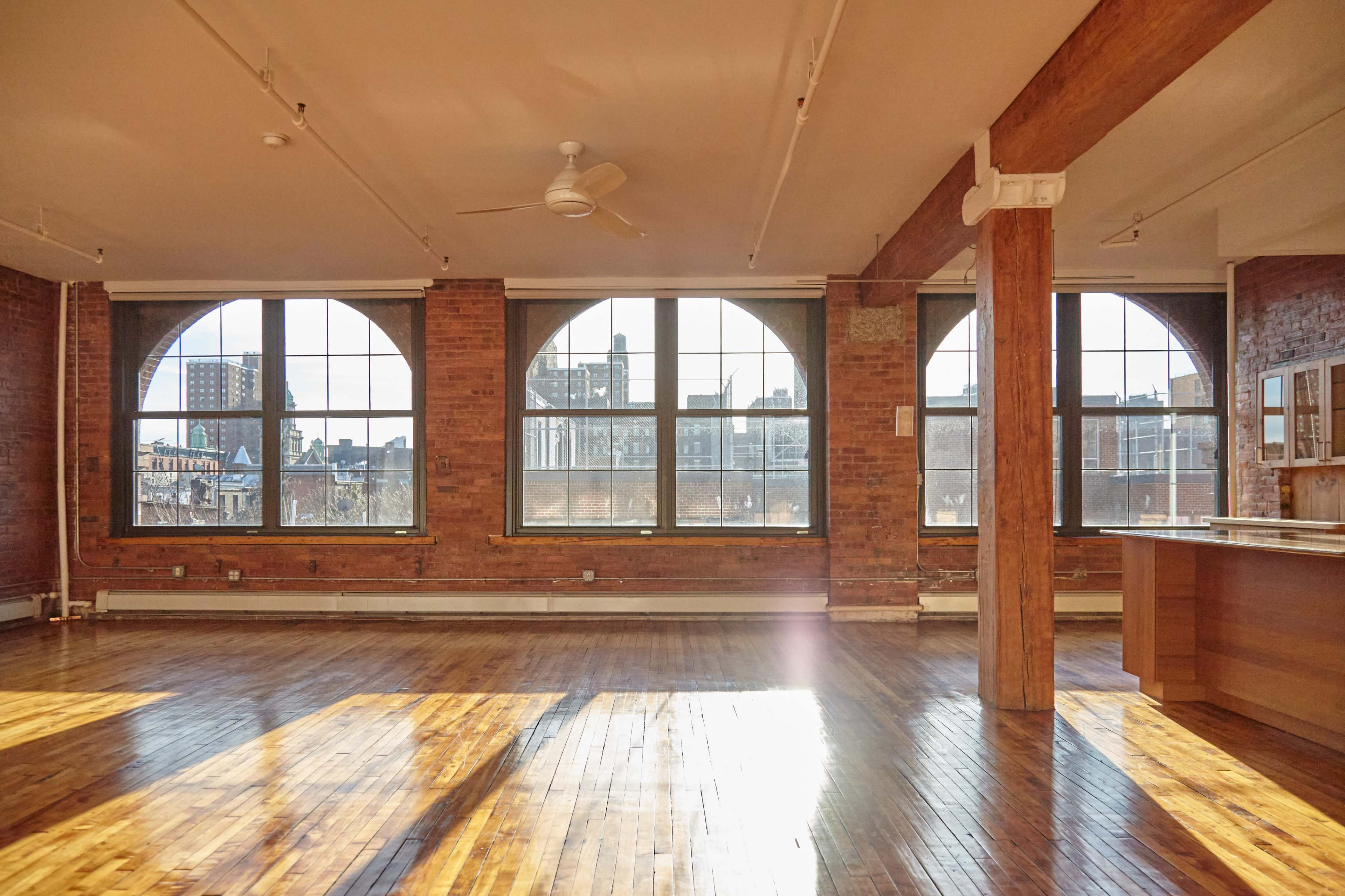 A spacious, well-lit interior with large windows, exposed brick walls, wooden floors, and a ceiling fan.
