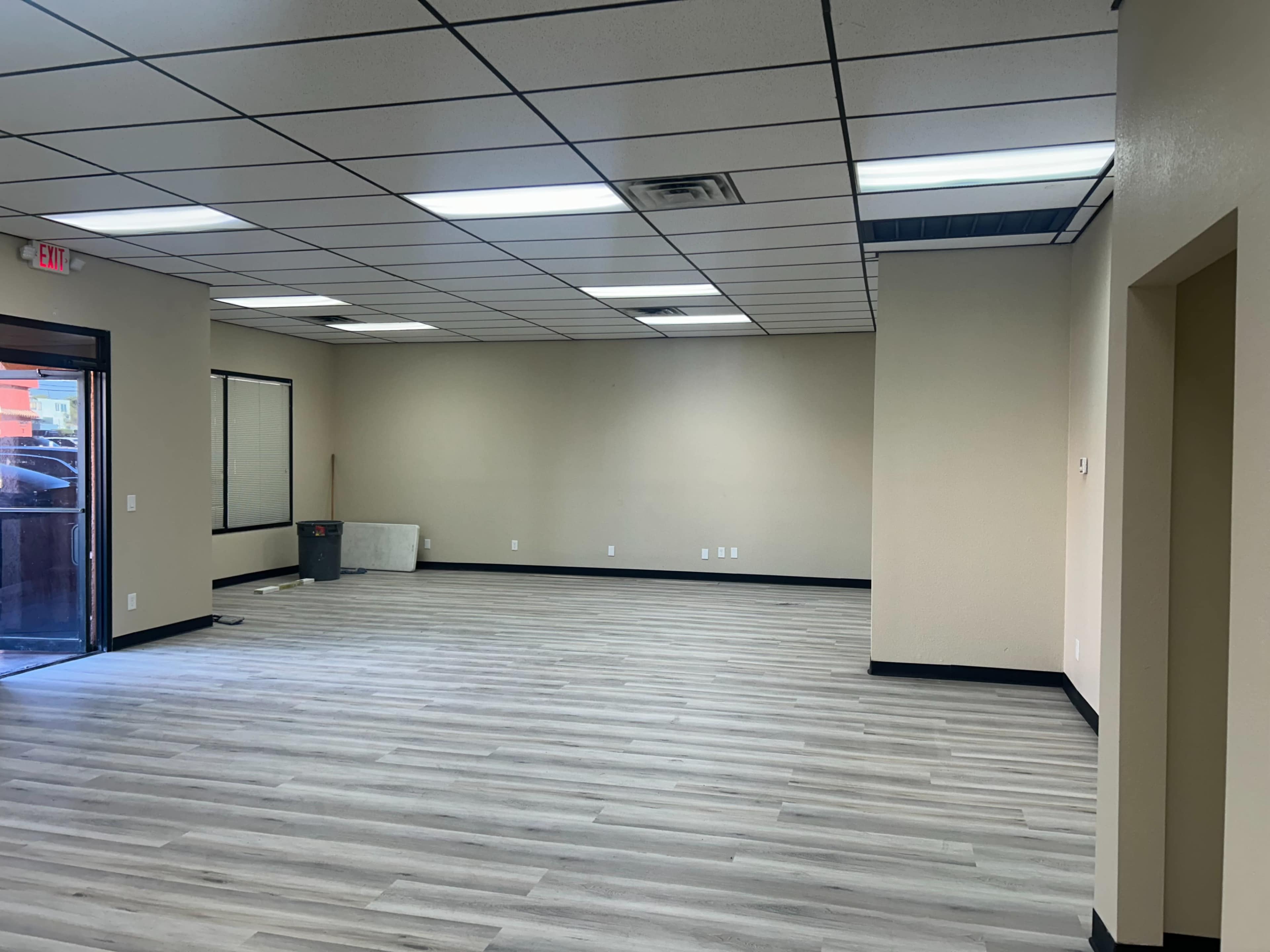 The image shows an empty office space with light-colored walls, a gray laminate floor, and minimal furnishings.