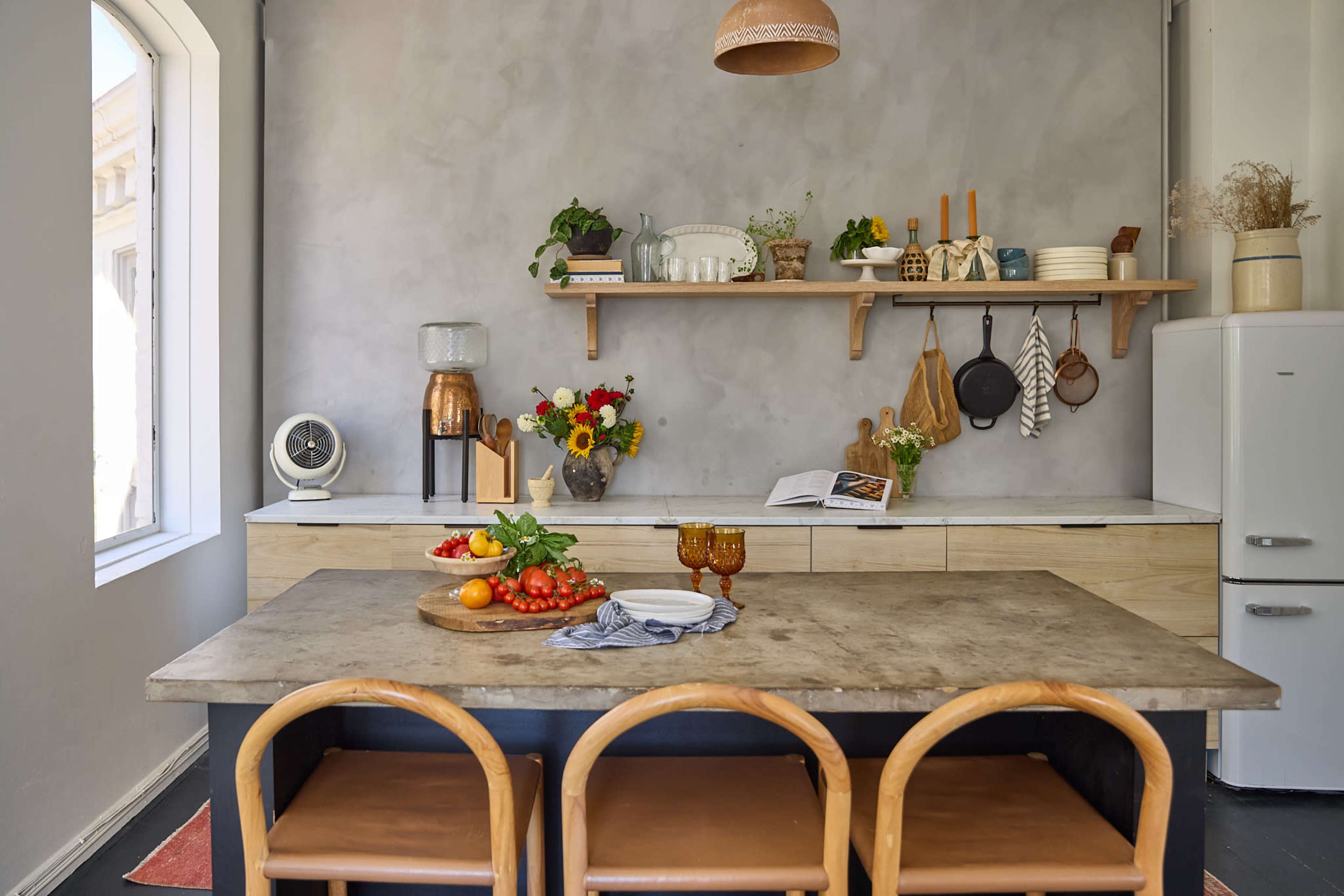 A modern kitchen features a concrete countertop, wooden shelves with kitchenware, and a dining table set with fresh vegetables and fruits.