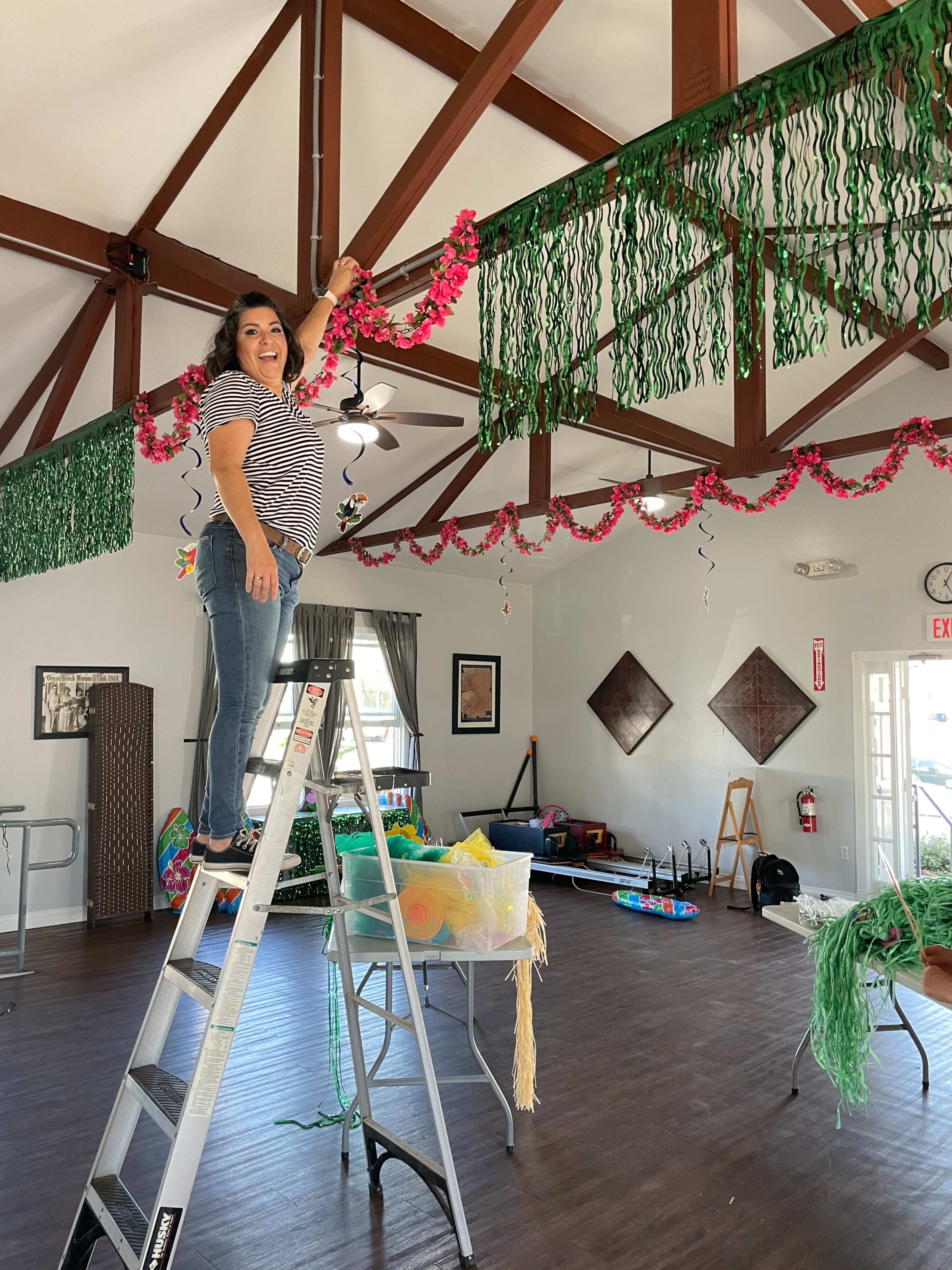 A woman stands on a ladder decorating the interior of a community hall with colorful garlands and greenery.