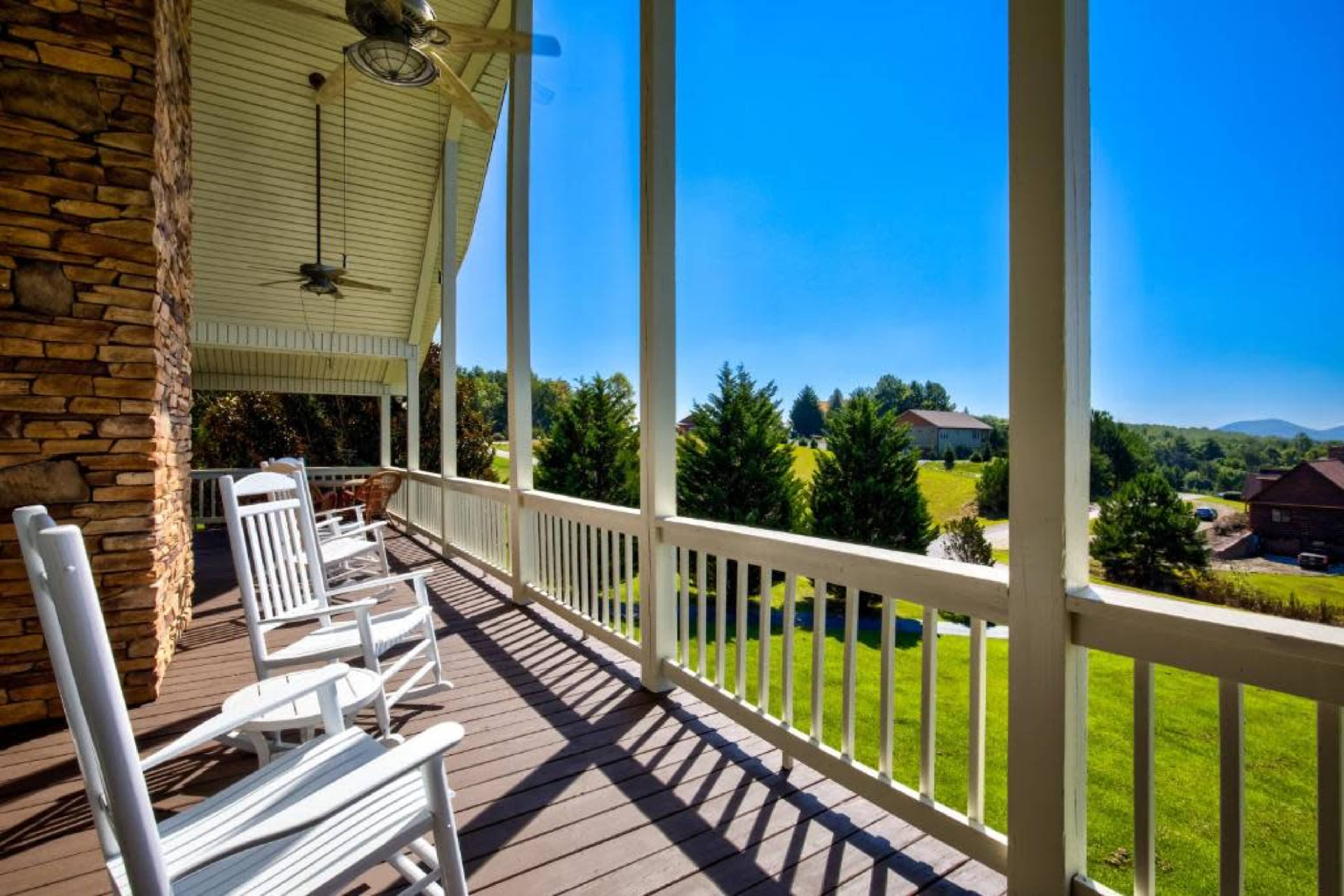 A wooden porch with rocking chairs overlooks a green landscape and distant hills under a clear blue sky.