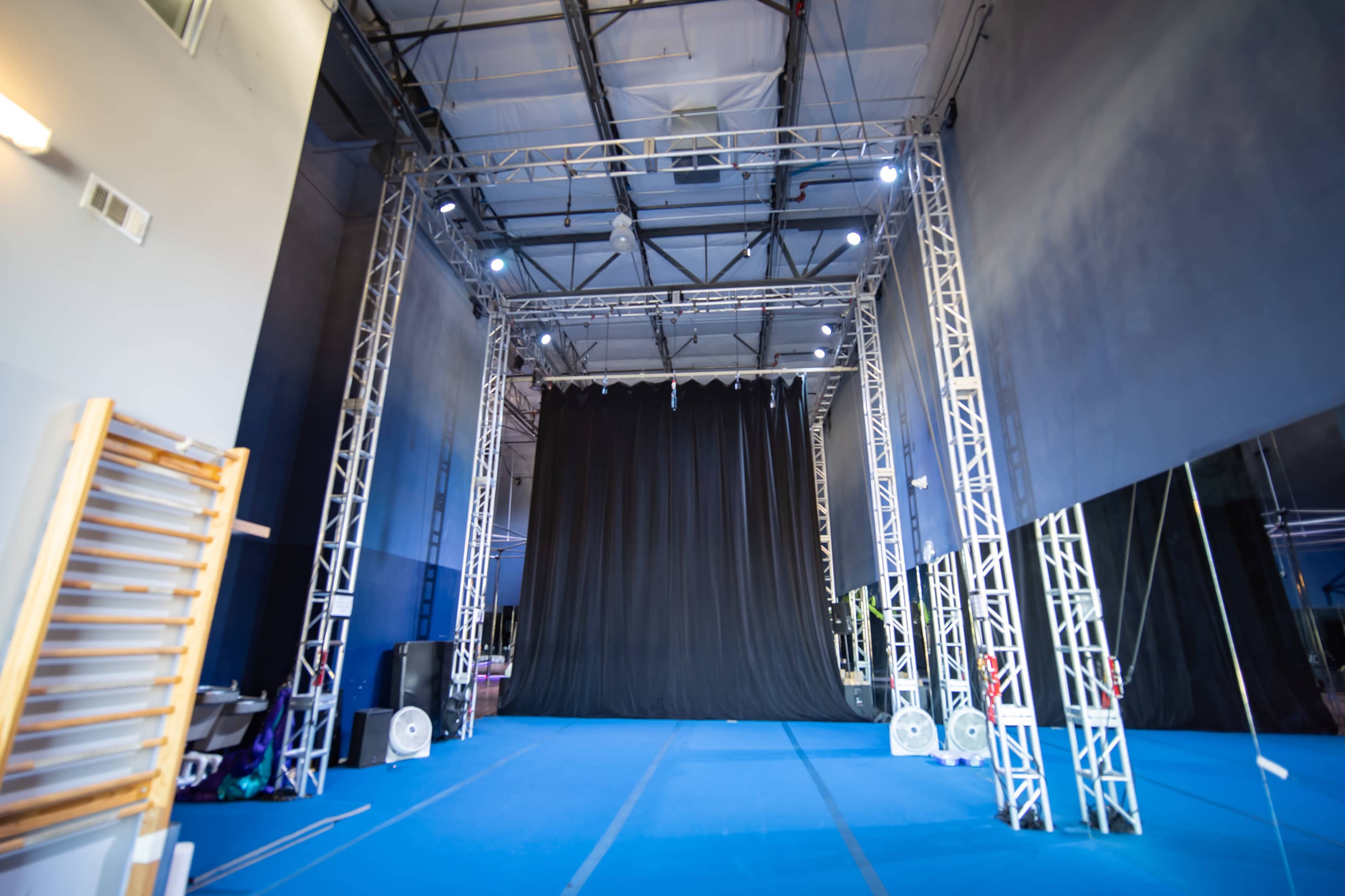 A large performance space with a black curtain at the back, surrounded by metal trusses and a blue floor.