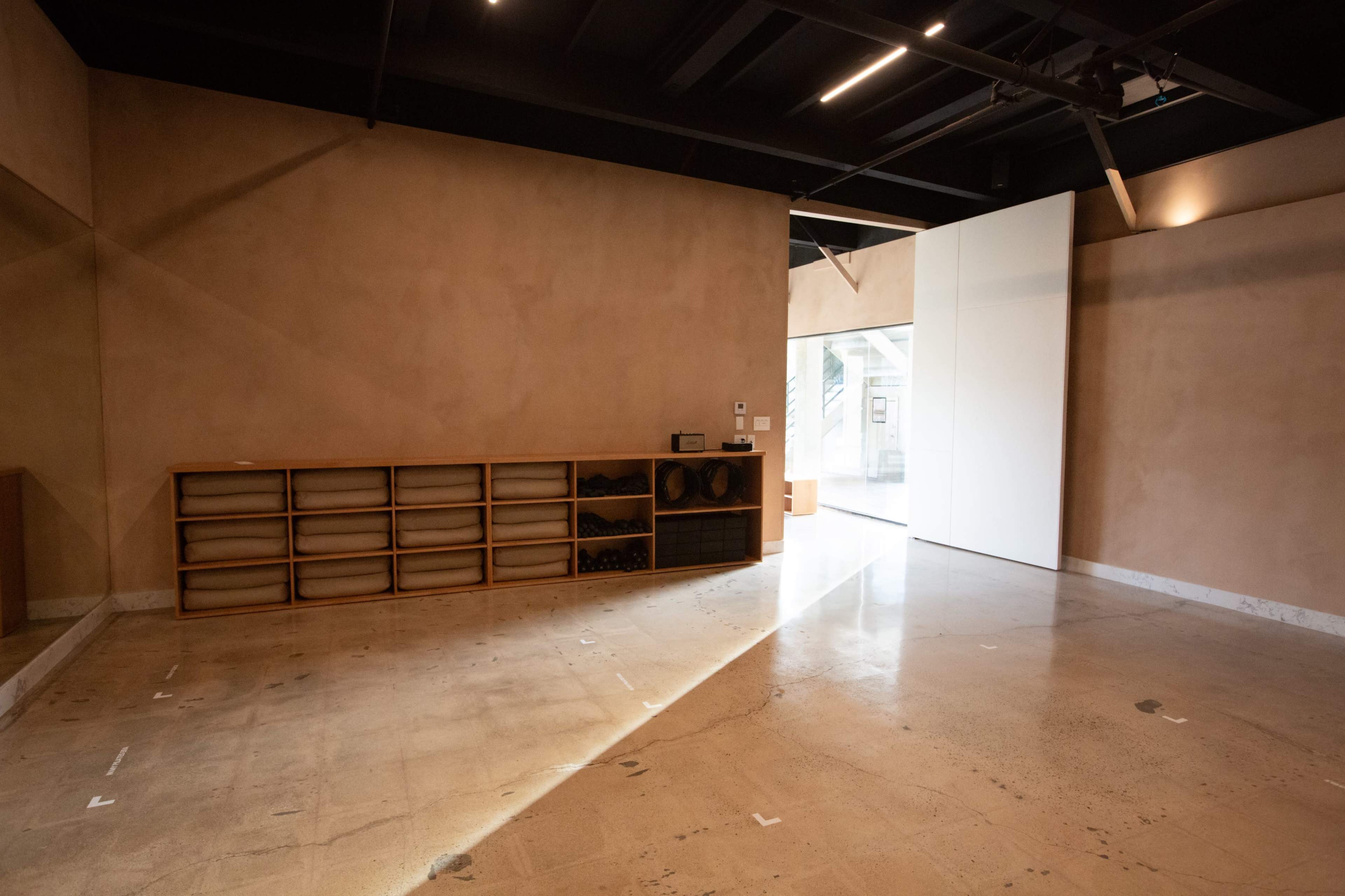 The image shows a minimalist interior space with a light-colored wall, a polished floor, and a series of wooden shelves along one side.