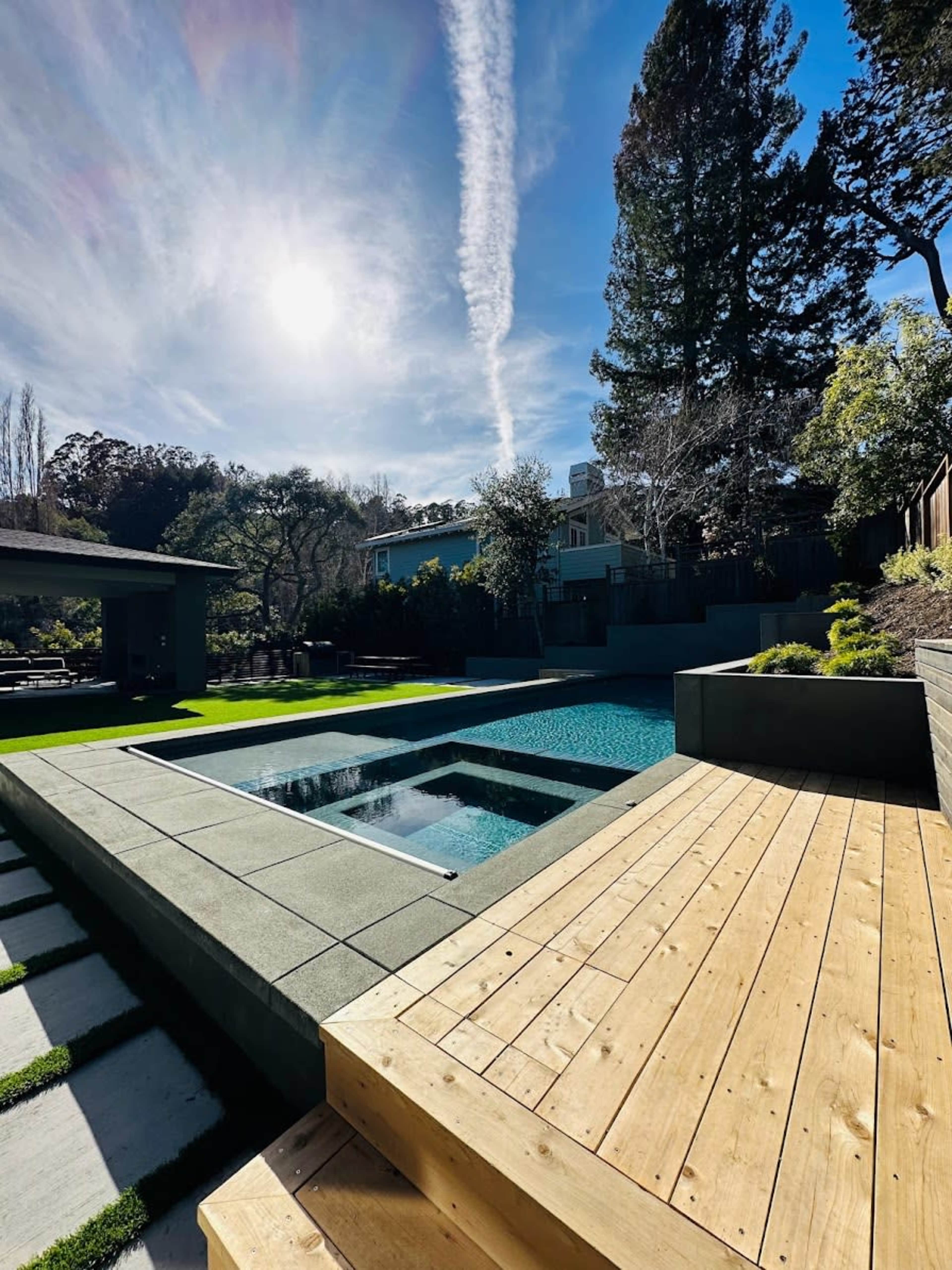 A modern backyard with a swimming pool surrounded by a stone patio, wooden deck, and lush greenery under a clear blue sky.