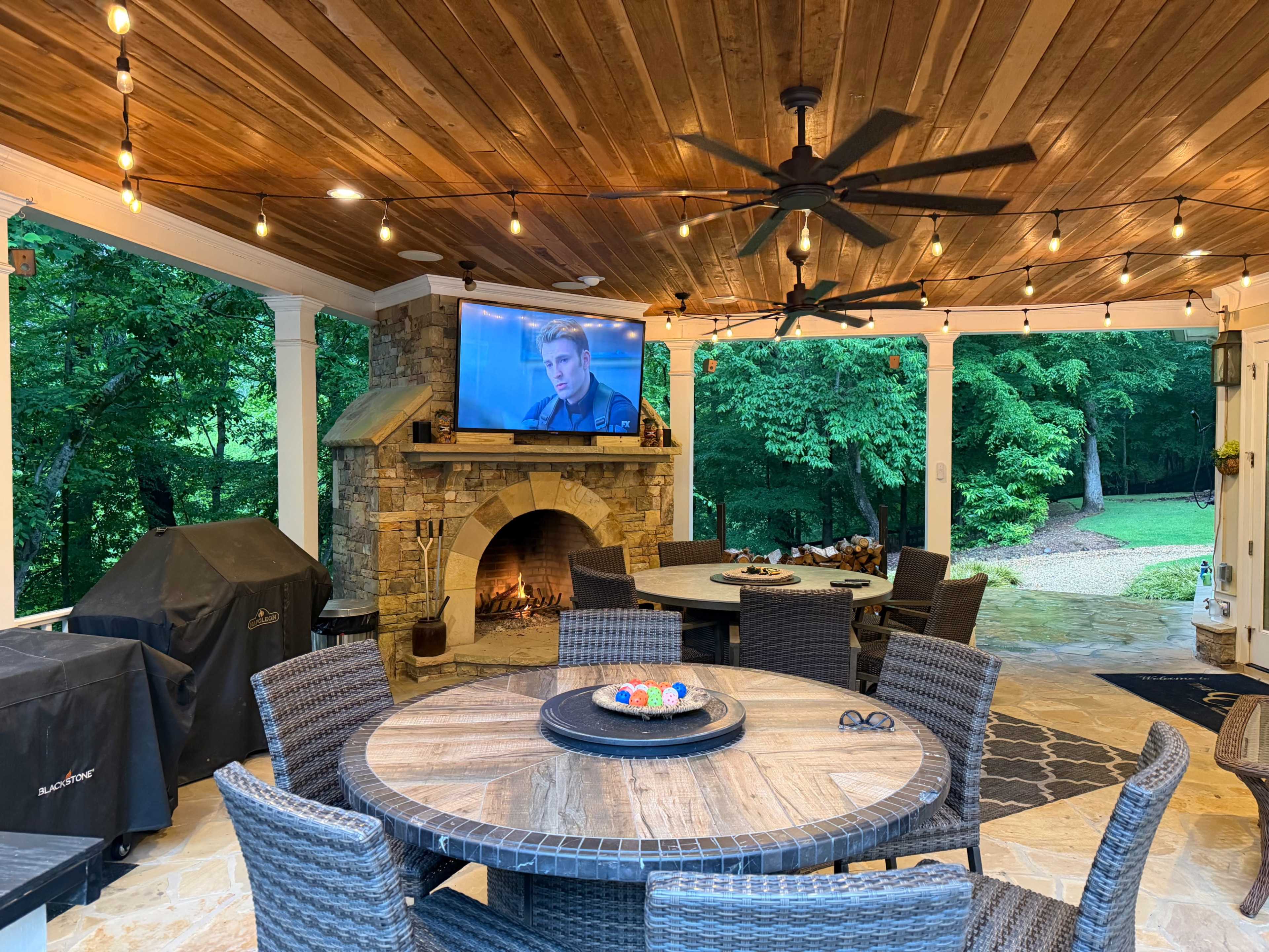 The image shows a covered outdoor patio featuring a round dining table, a large stone fireplace, and a television mounted on the wall, with string lights overhead and greenery visible in the background.