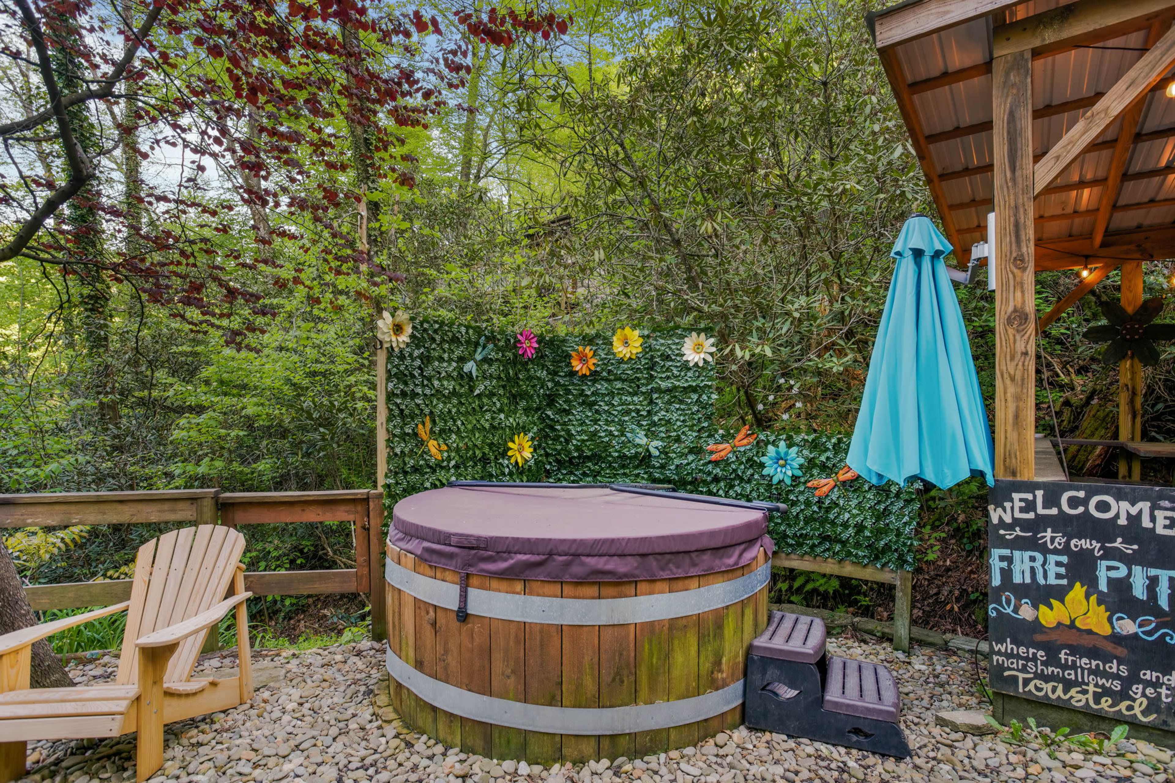 A wooden hot tub surrounded by greenery, with a seating area, a decorative sign, and a blue umbrella nearby.