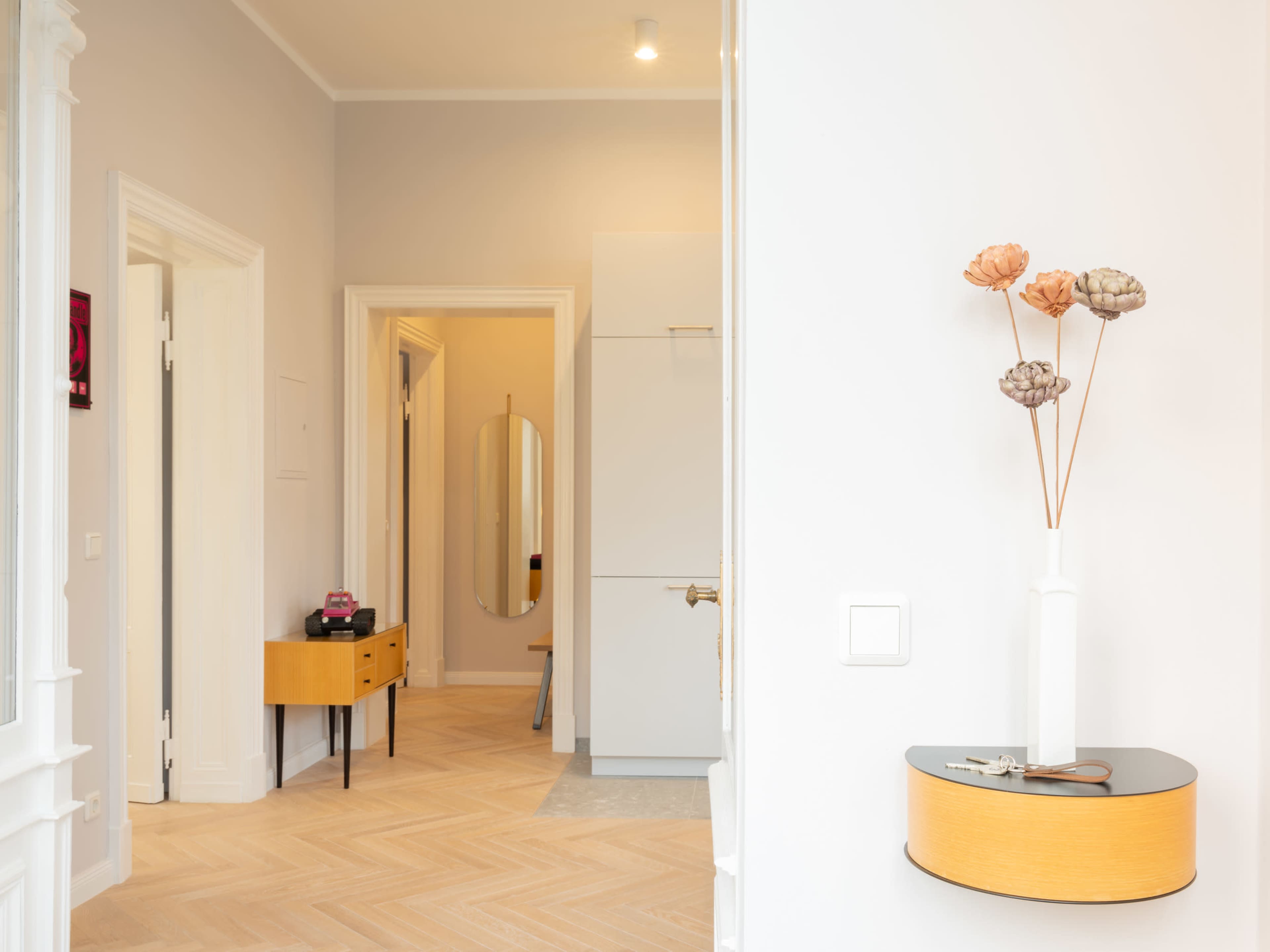 A hallway features light-colored walls, a wooden floor with a herringbone pattern, and a decorative console table with a vase of flowers.