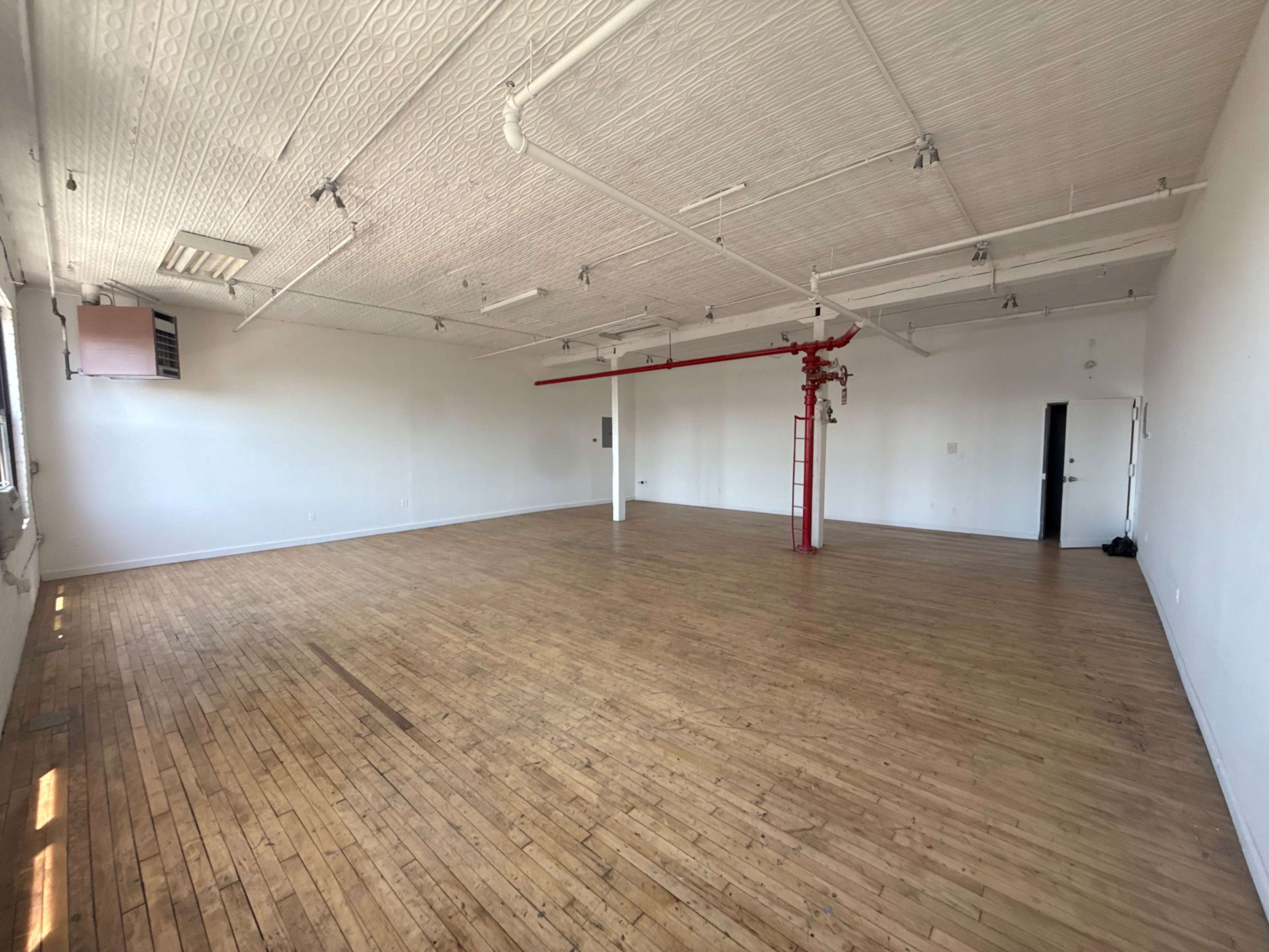 The image shows a spacious, empty room with hardwood floors and white walls, featuring exposed ceiling details and a red pipe running along the ceiling.