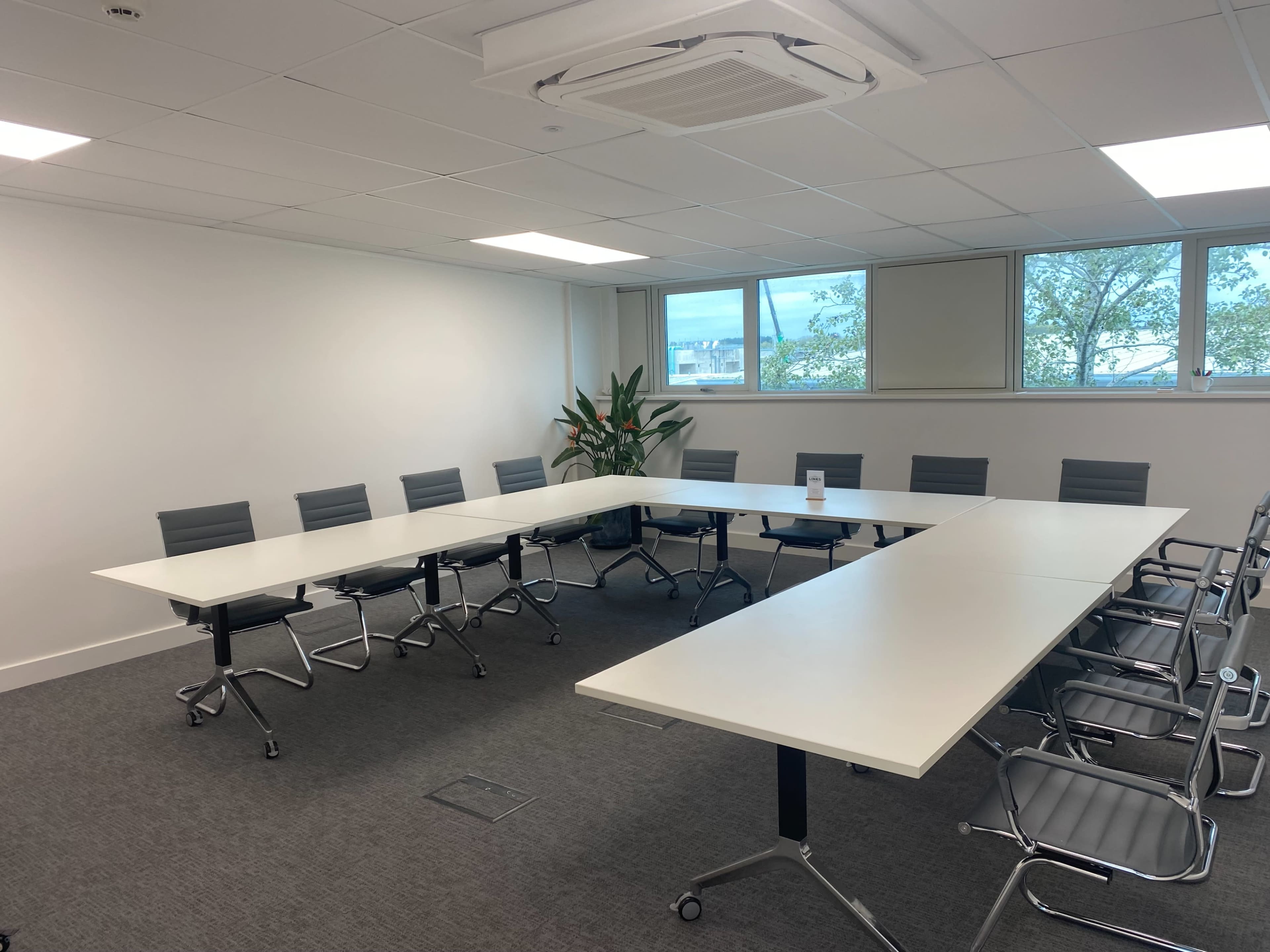 A conference room features a large, white rectangular table surrounded by multiple gray chairs, with windows showing greenery outside.