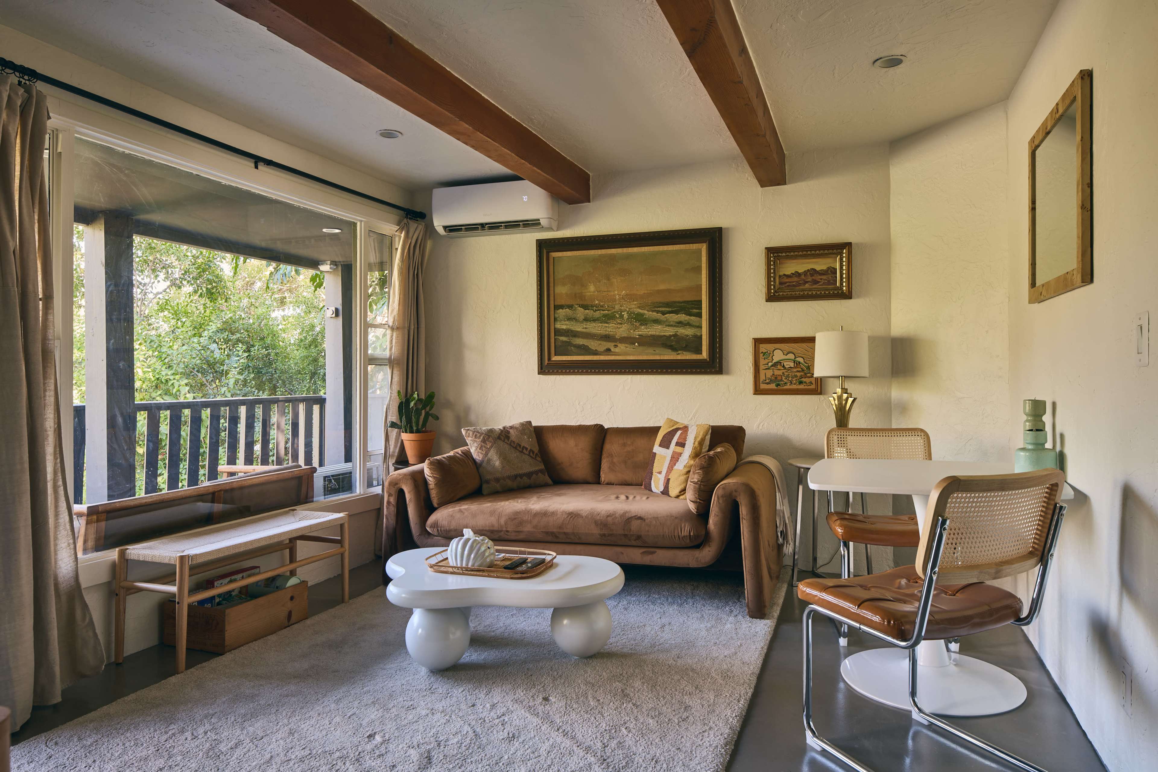 The image shows a modern living room with wooden beams, a brown couch, a white table and chairs, and large windows looking out onto greenery.
