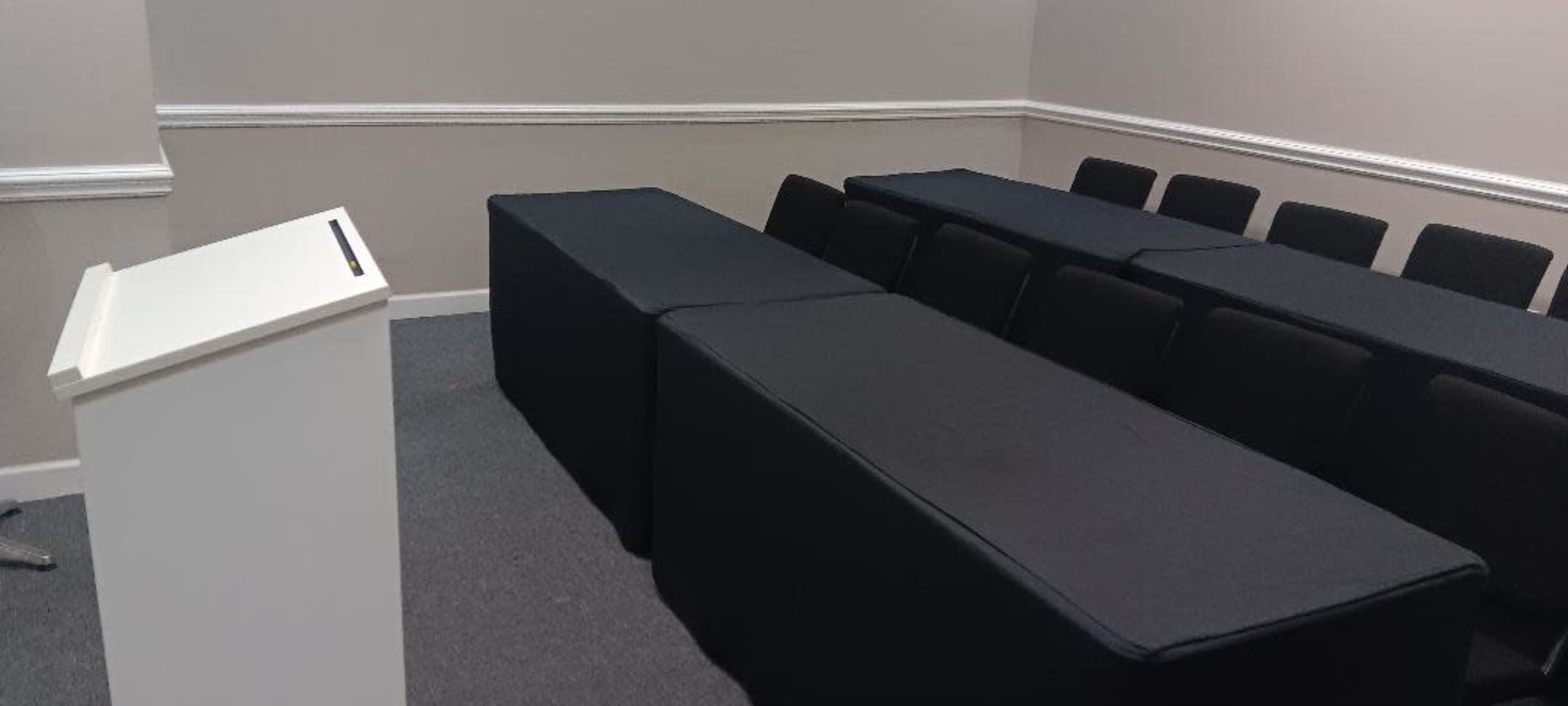 A conference room with two long tables covered in black cloth and several chairs arranged behind them, alongside a white podium at the front.