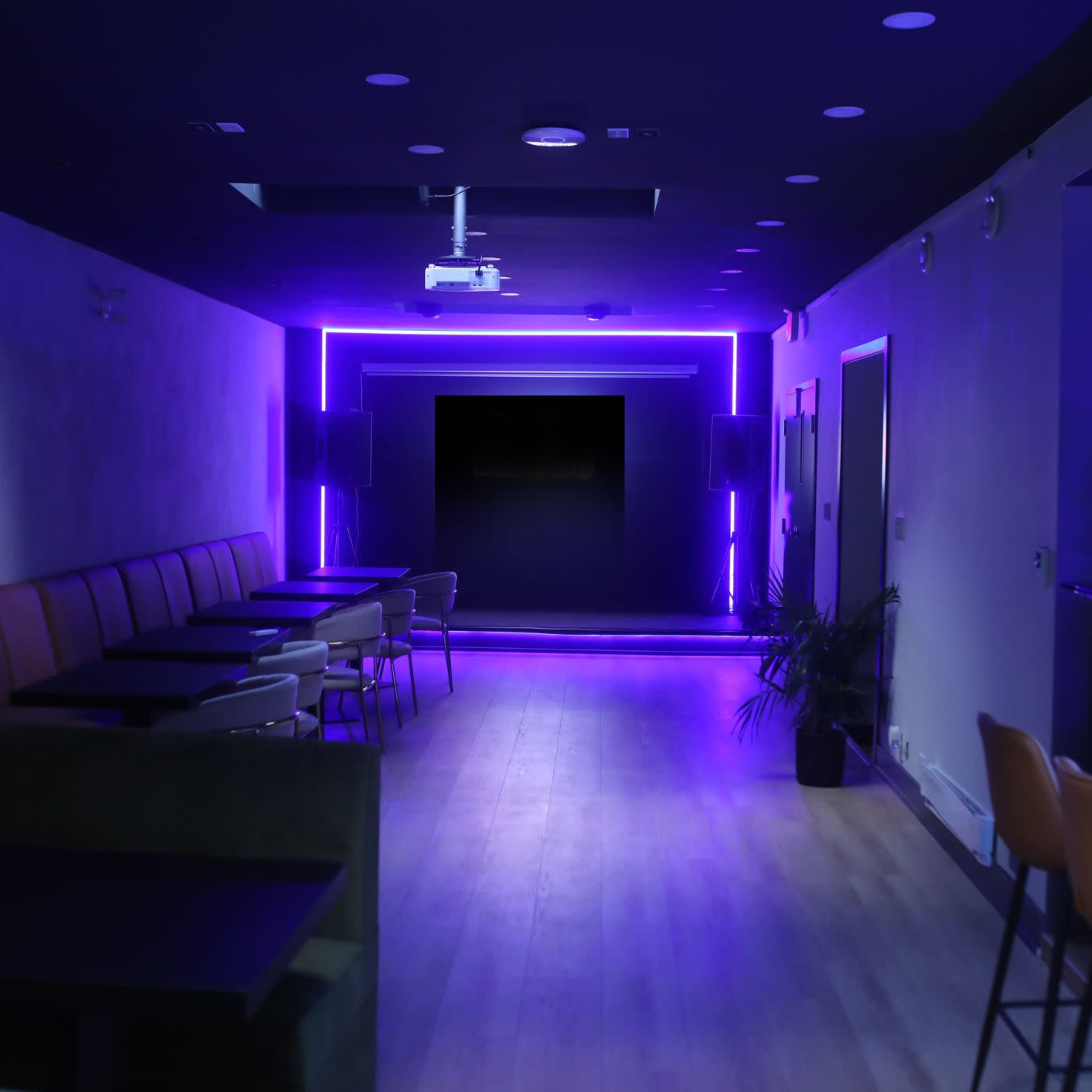 A dimly lit corridor leads to a space illuminated by purple neon lights, with tables arranged on either side.