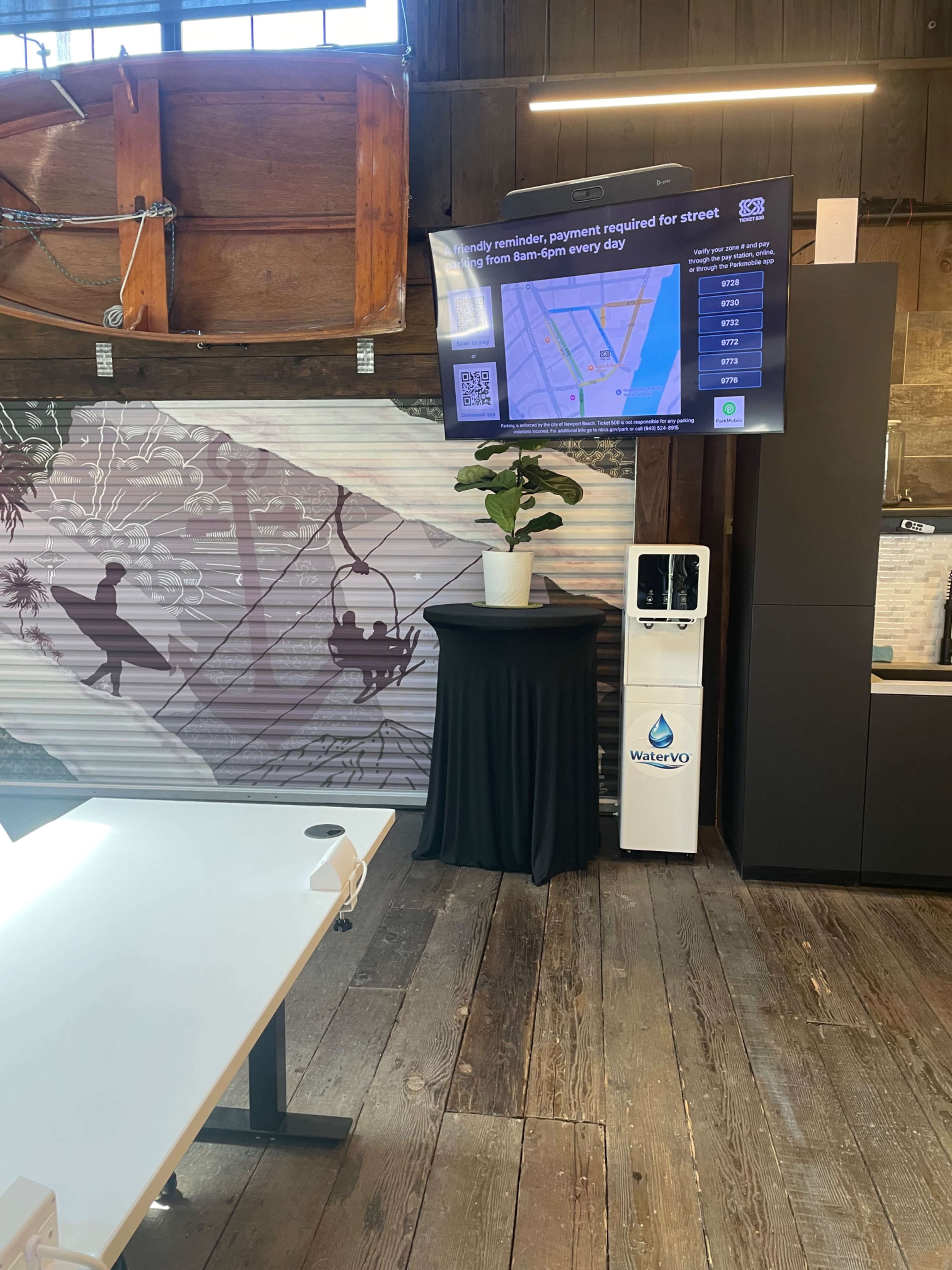 The image shows a digital display mounted on a wooden wall, featuring a map and a reminder about street parking payment, alongside a water cooler and a plant on a table.