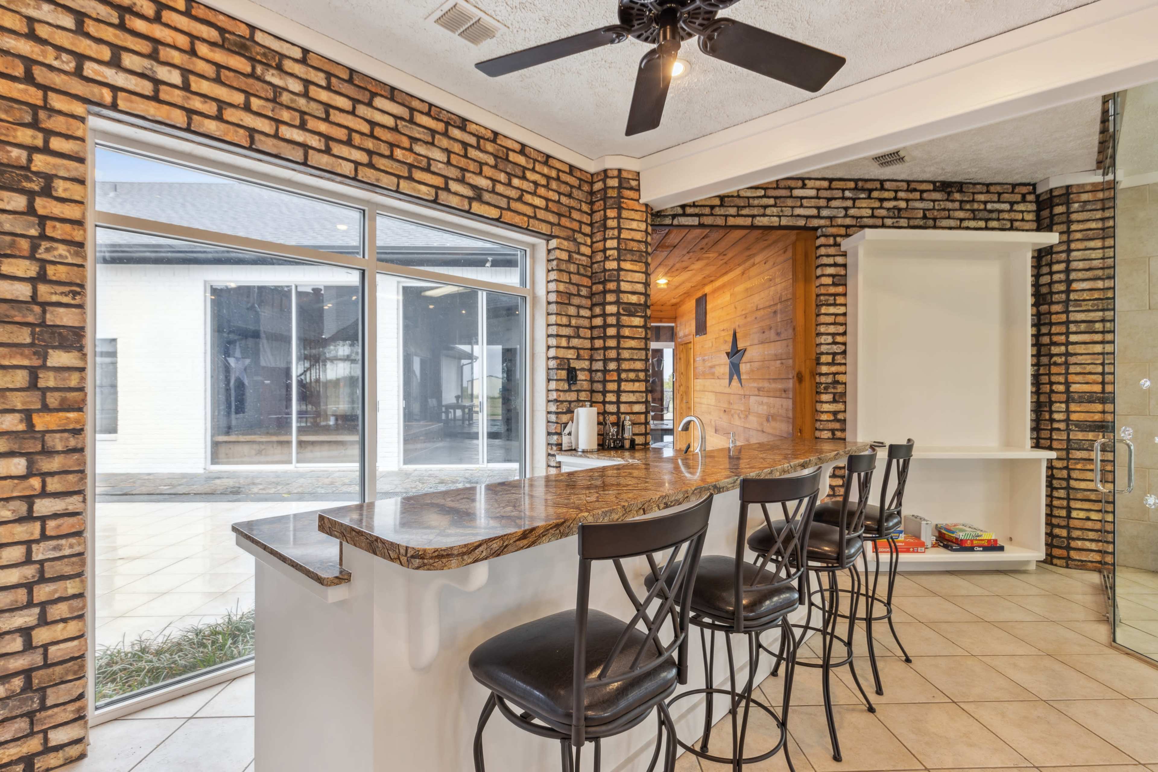 The image shows a kitchen with a granite countertop and bar seating, featuring brick walls and large windows that overlook an outdoor area.