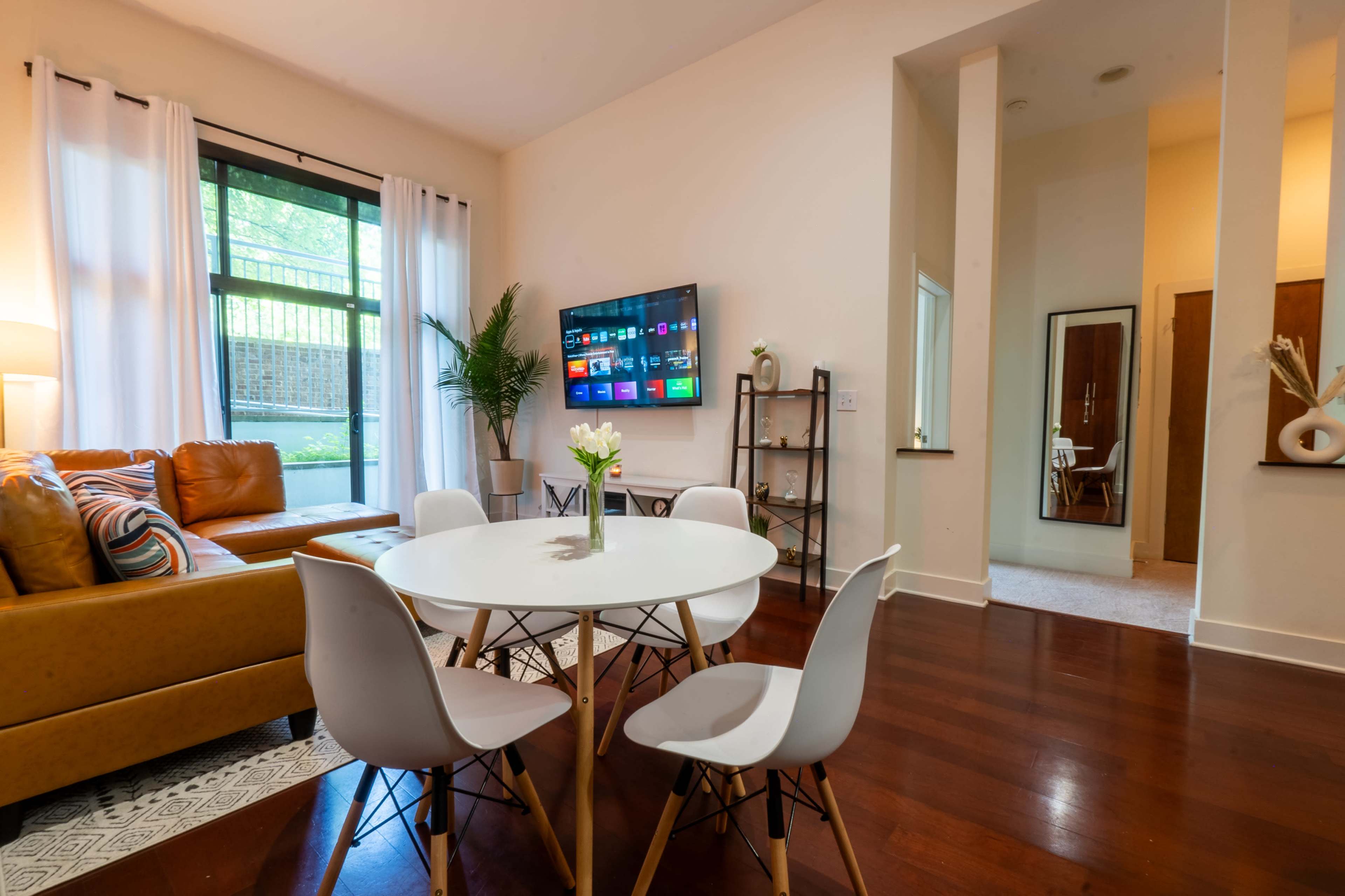 A modern living space featuring a yellow sofa, a round dining table with white chairs, and a wall-mounted television.