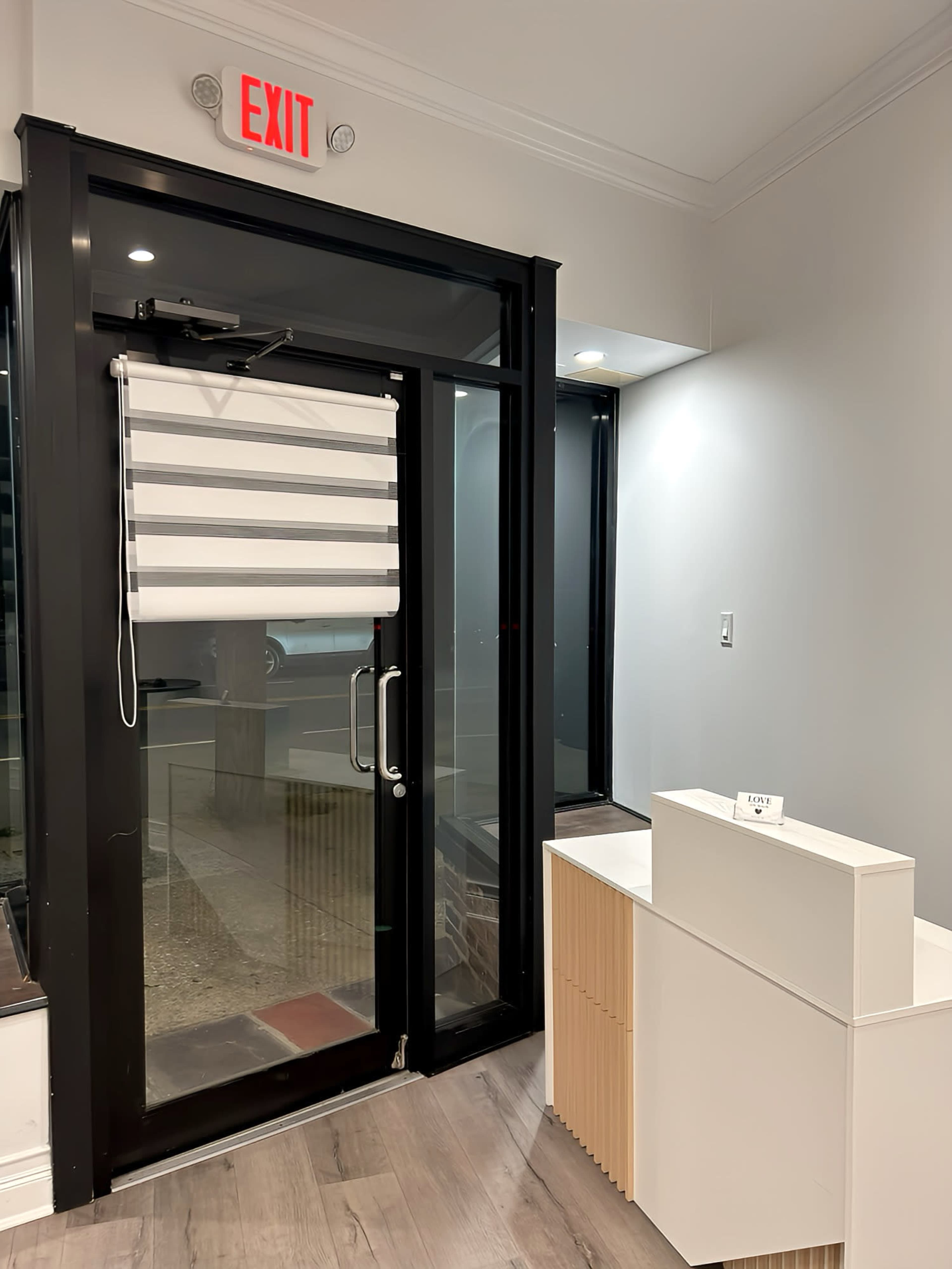 The image shows a modern entryway with a glass door, a roll-up blind, and a reception desk in front of the entrance.