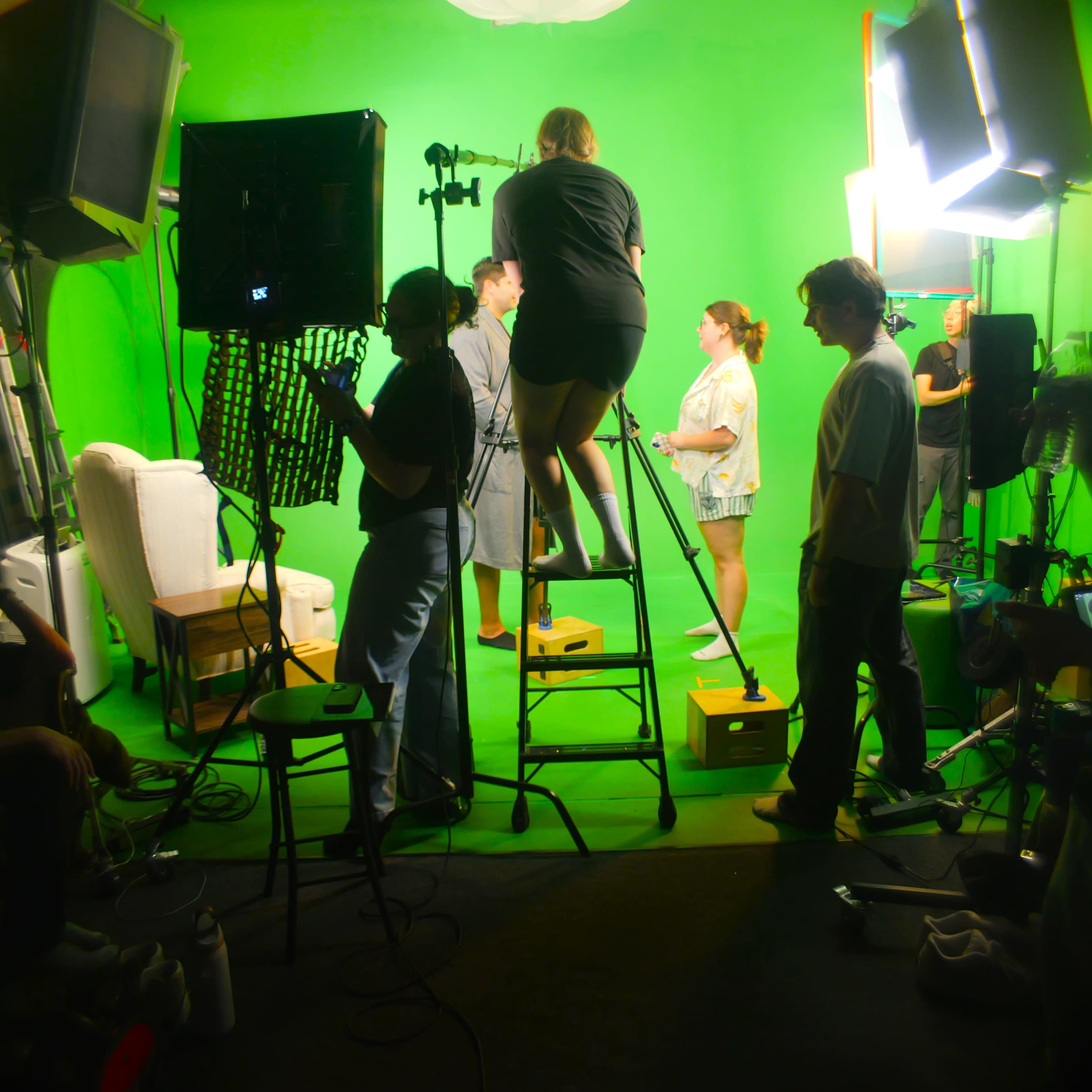 A film crew is working in a studio with a green screen backdrop, while some members adjust equipment and others interact on set.