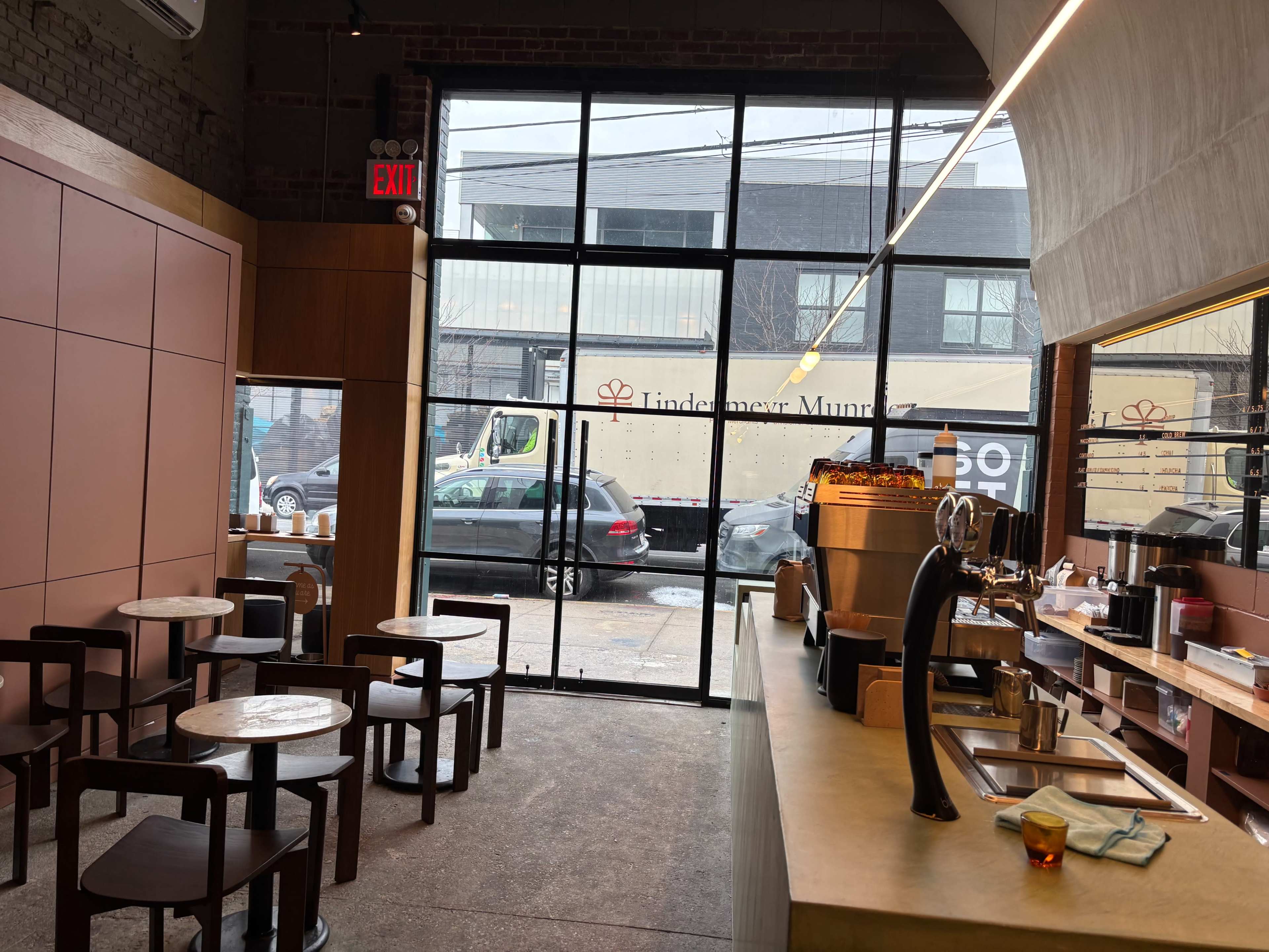The image shows the interior of a modern café with wooden furnishings, small round tables, and a counter displaying coffee equipment, while large glass windows provide a view of the street outside.