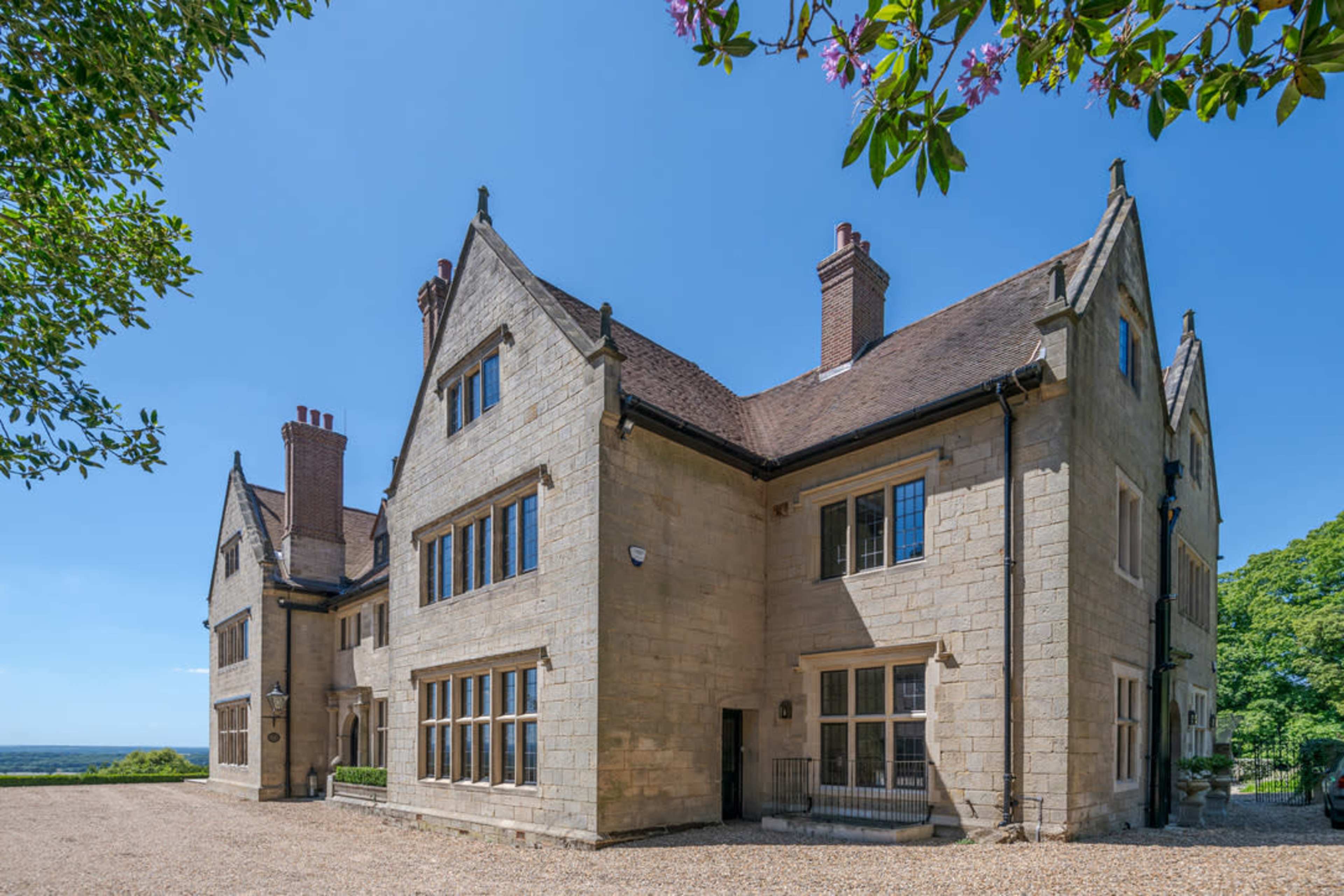 The image shows a large stone house with multiple chimneys and large windows, set against a clear blue sky.