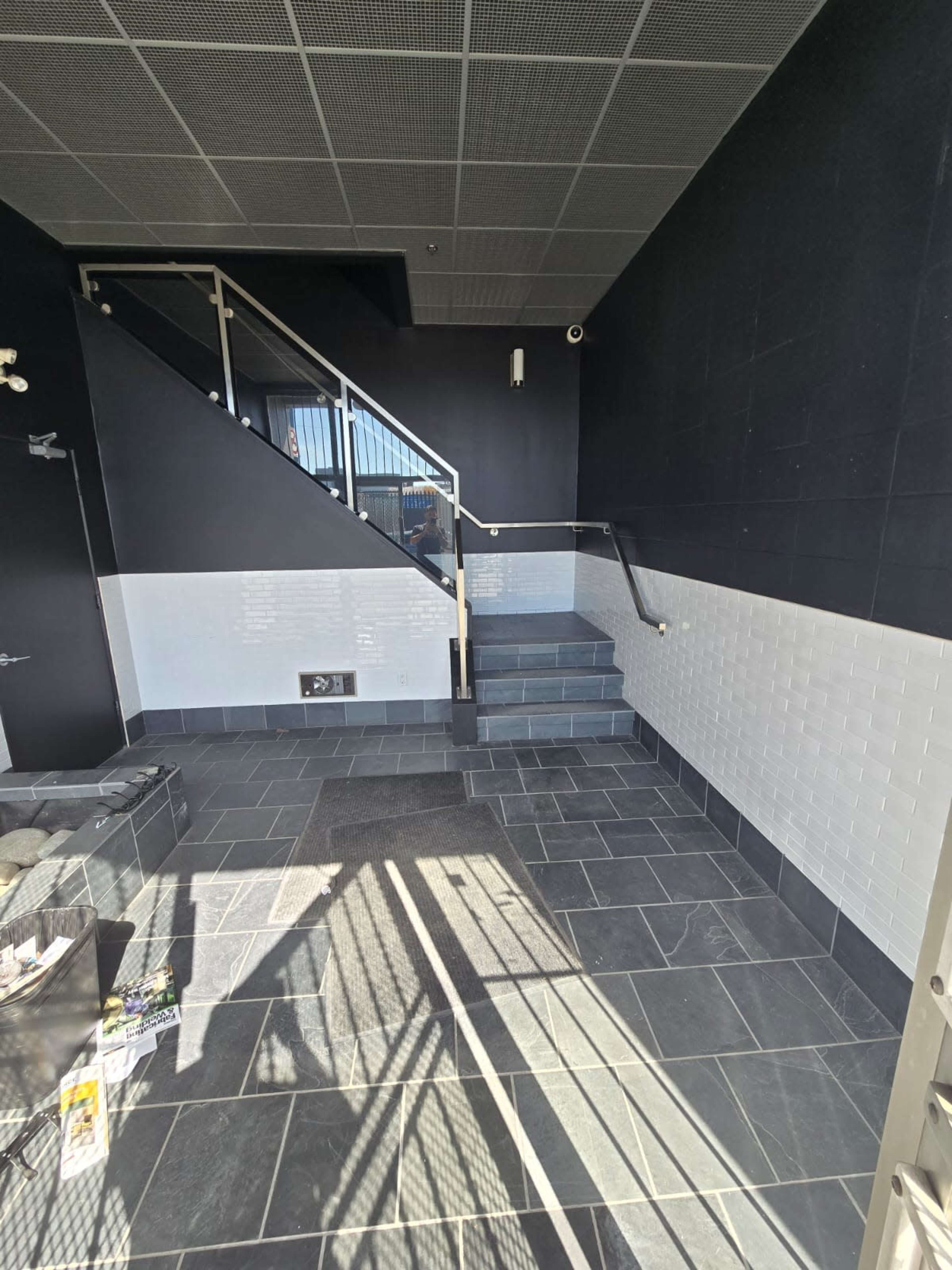 The image shows a modern entrance area with a dark tiled floor, a staircase leading upward, and a white tiled wall section.