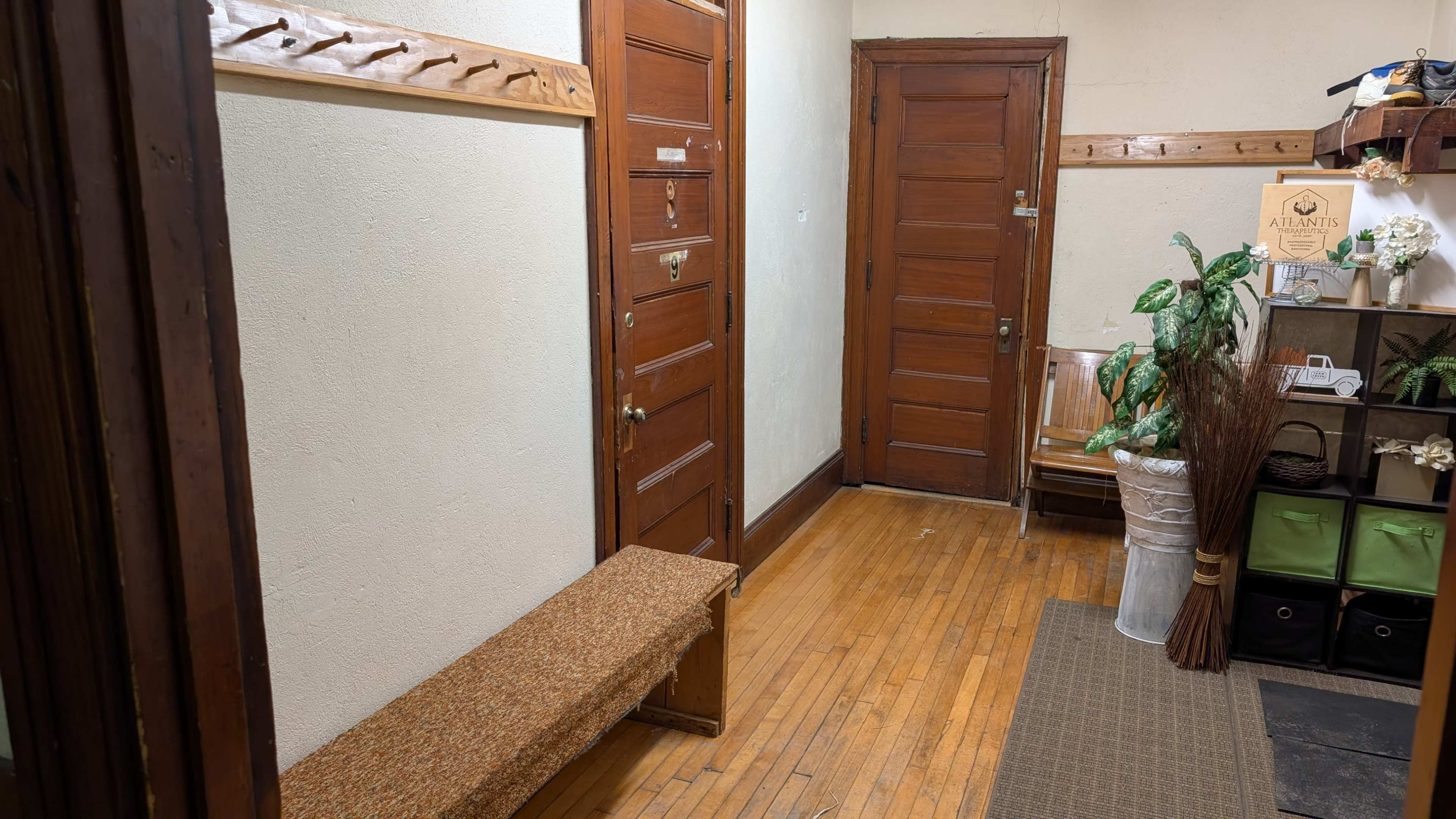 The image shows a hallway with wooden floors, two doors, a bench, a potted plant, and a storage unit with green fabric bins.