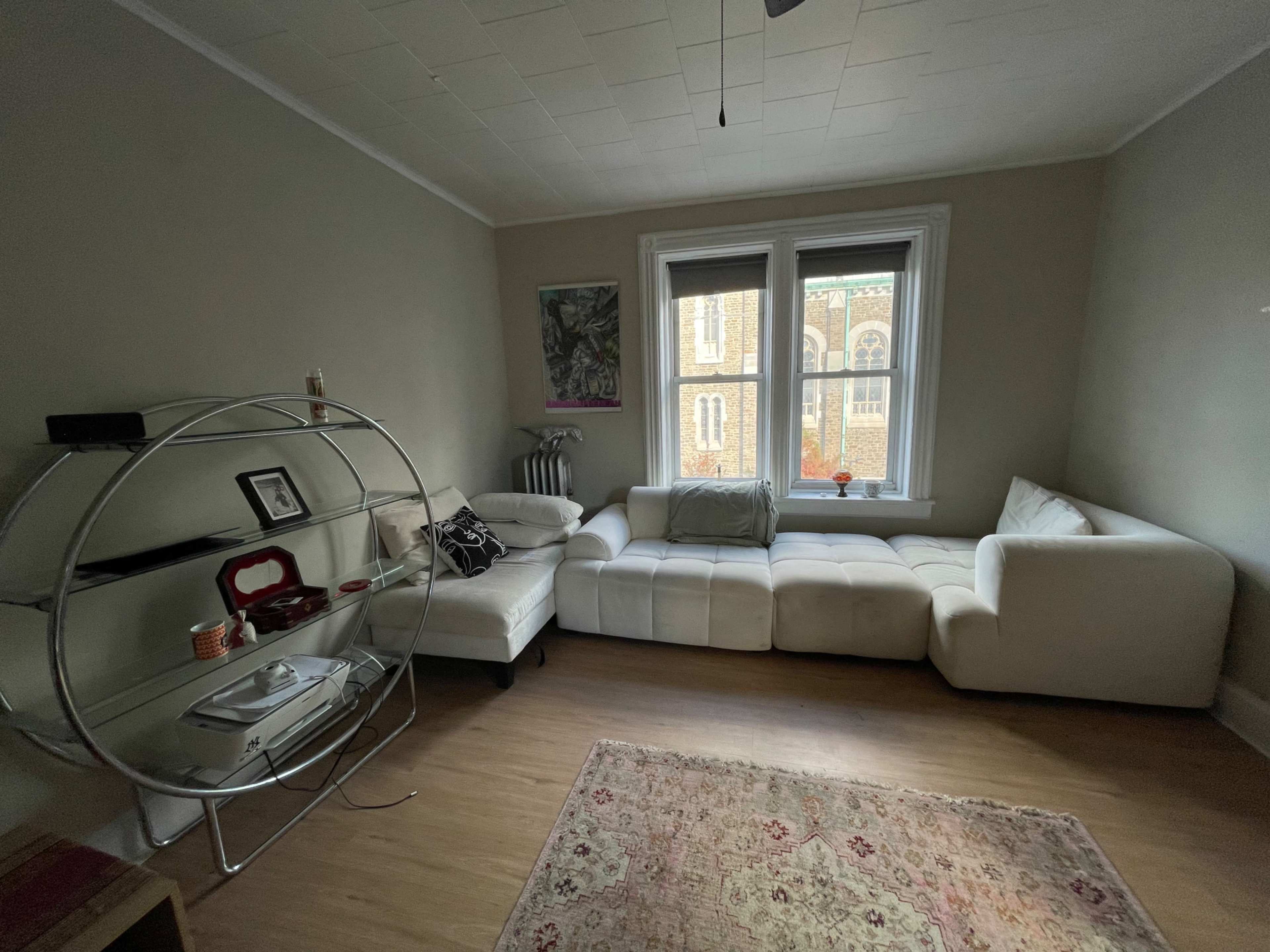 The image shows a minimalist living room with a white sectional couch, a circular metal shelf, and a large window facing an exterior view.