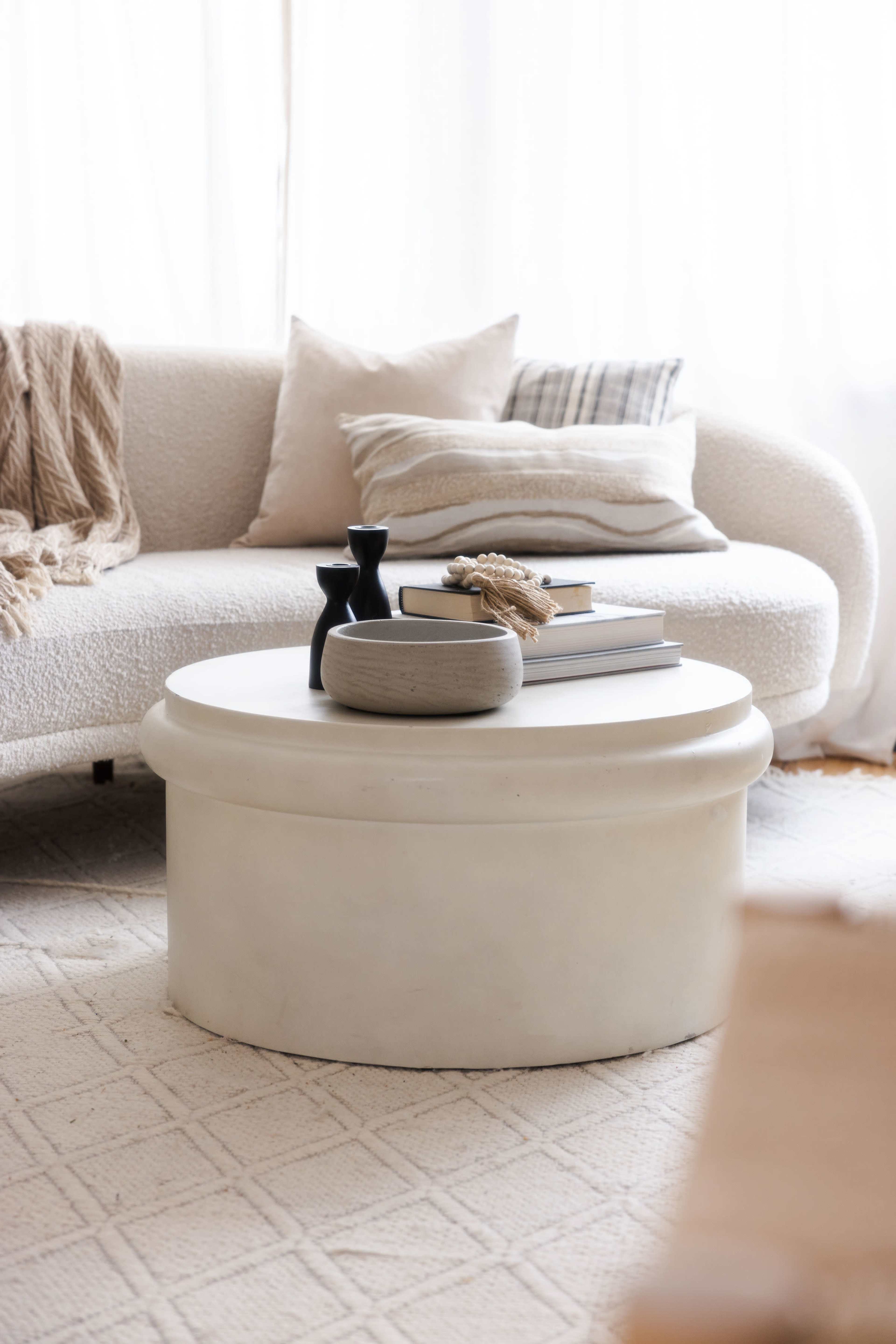 A light-colored, round coffee table is topped with a stack of books, a decorative bowl, and two vases, positioned in a cozy living room with a textured sofa and soft textiles.