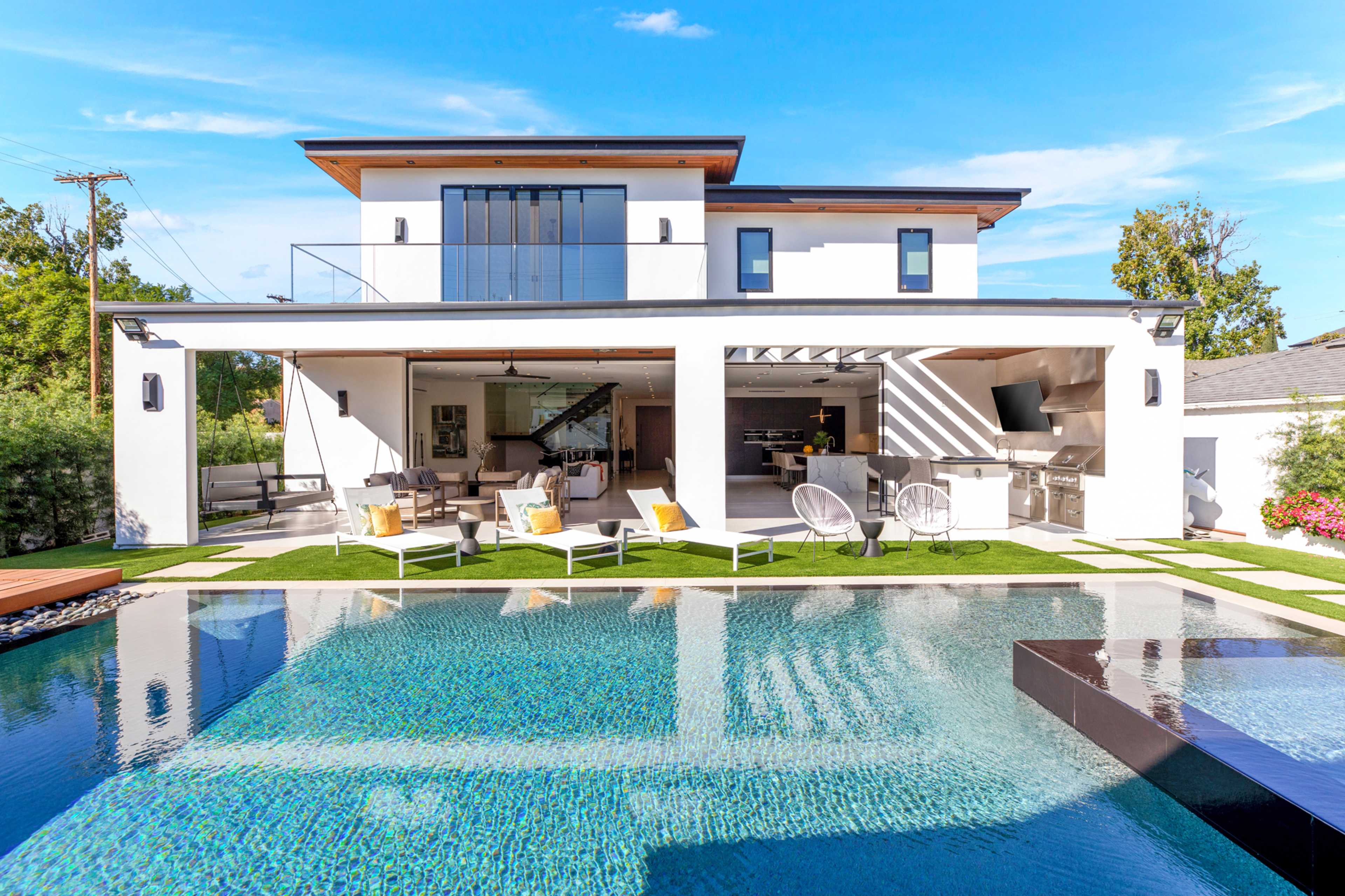 A modern two-story house features an outdoor patio with seating and a pool surrounded by landscaped greenery.
