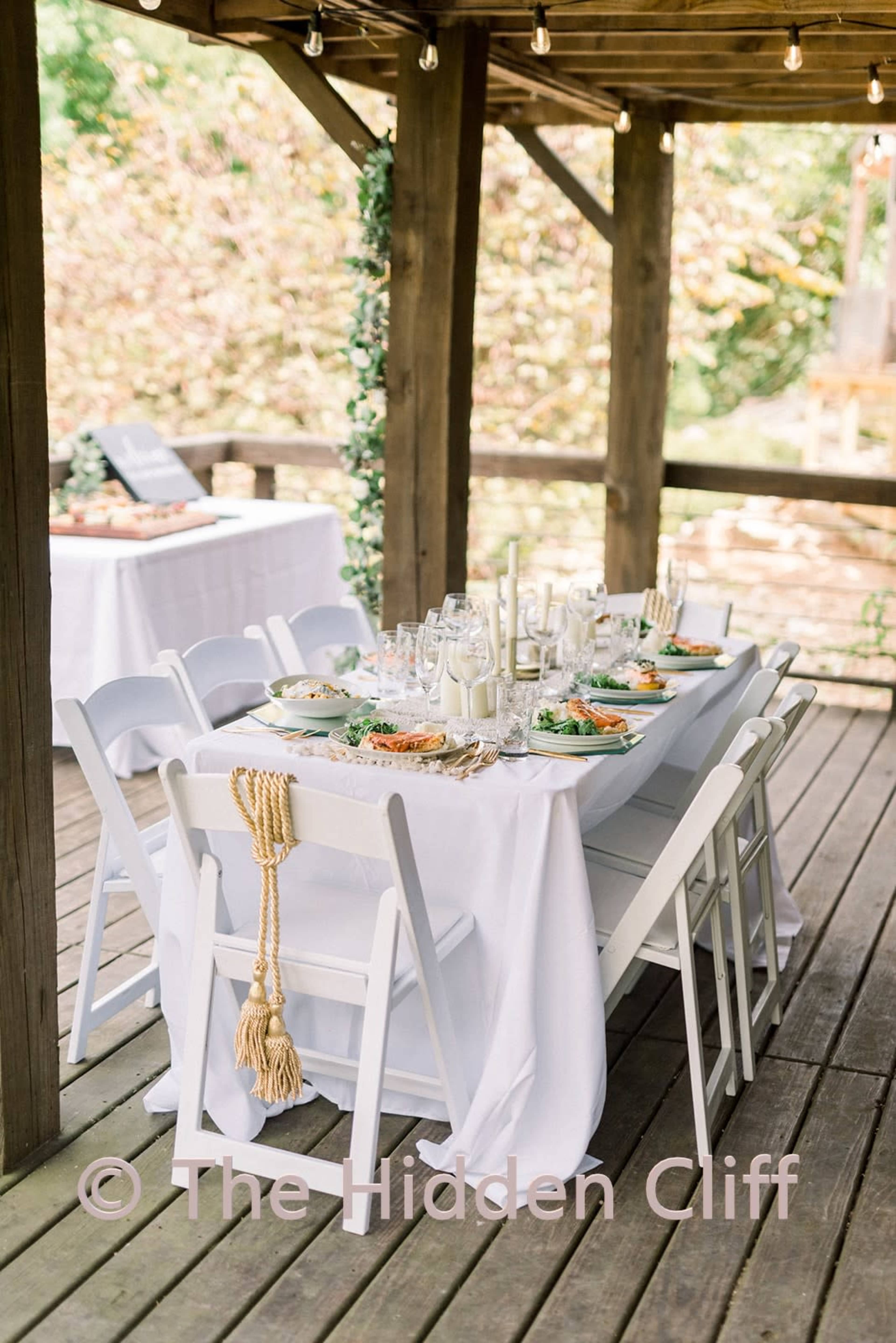A long dining table is set with plates of food and drinks under a wooden gazebo, surrounded by strings of lights and greenery.