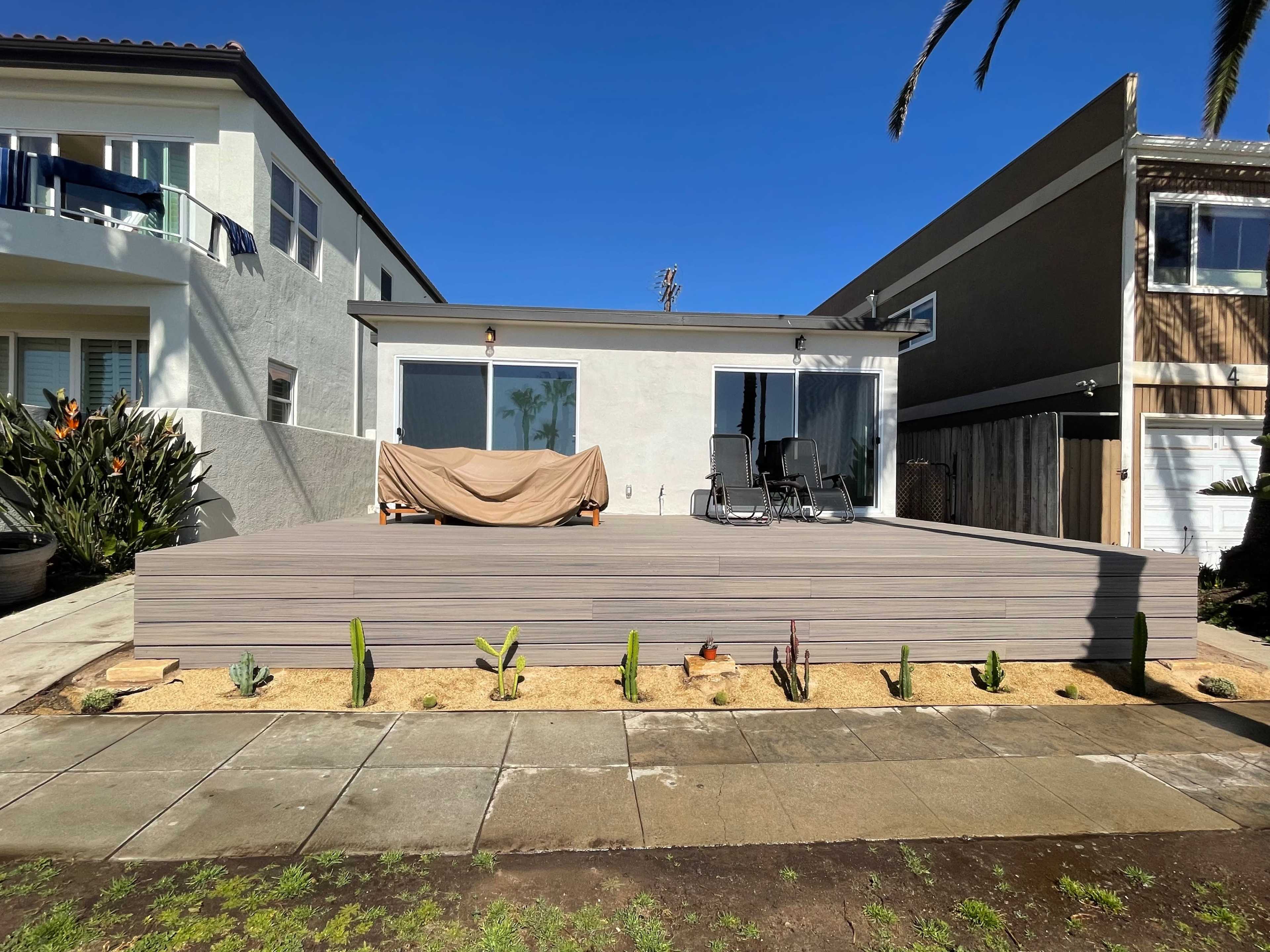 The image depicts a large wooden deck in front of a house, featuring two chairs and a covered area, with a landscaped garden of cacti in the foreground.
