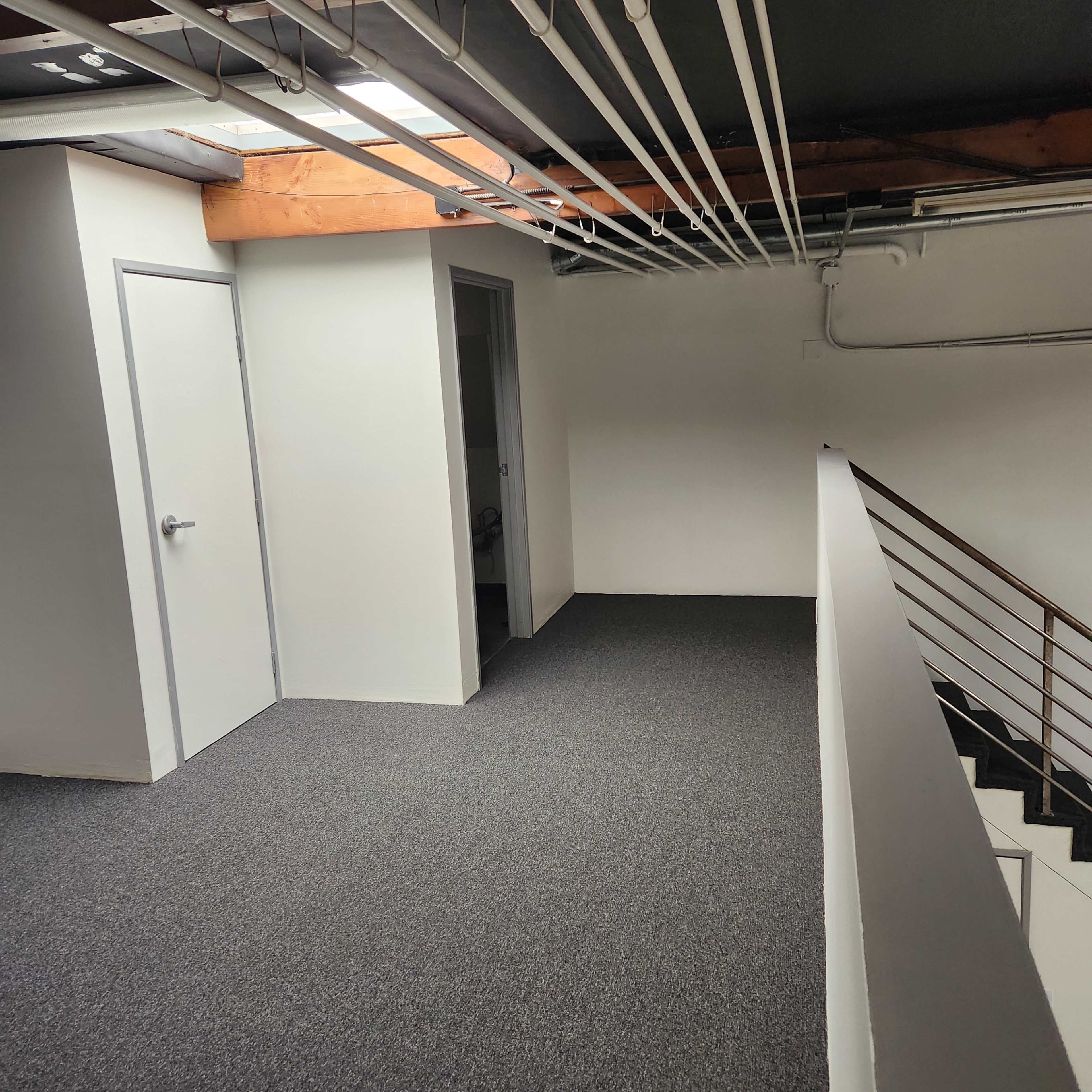 A carpeted hallway with two doors on the left and a staircase on the right, under a ceiling with exposed pipes.