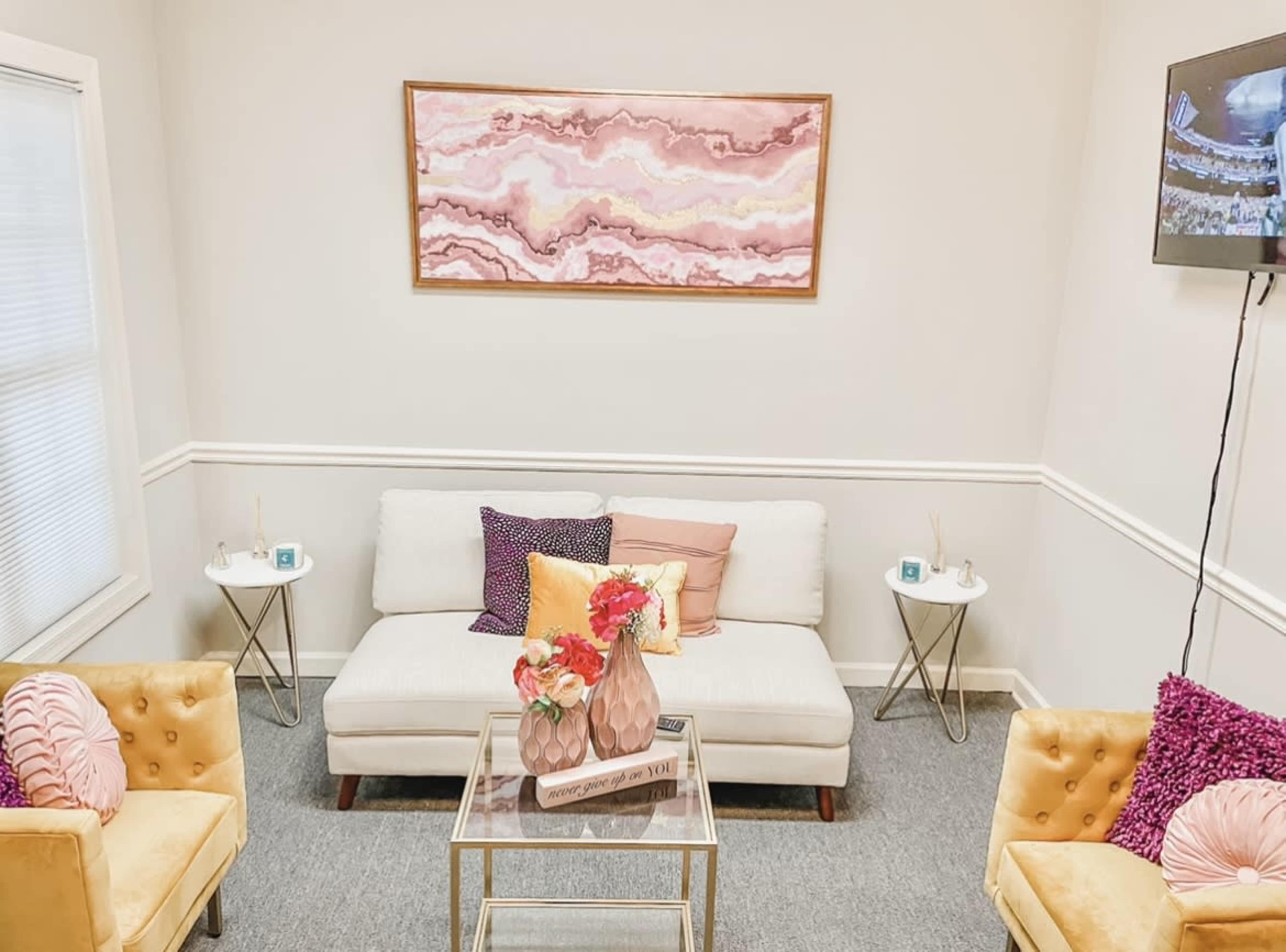 A small, elegantly arranged waiting area with a white sofa, two yellow chairs, a glass coffee table, and a large wall art piece above.