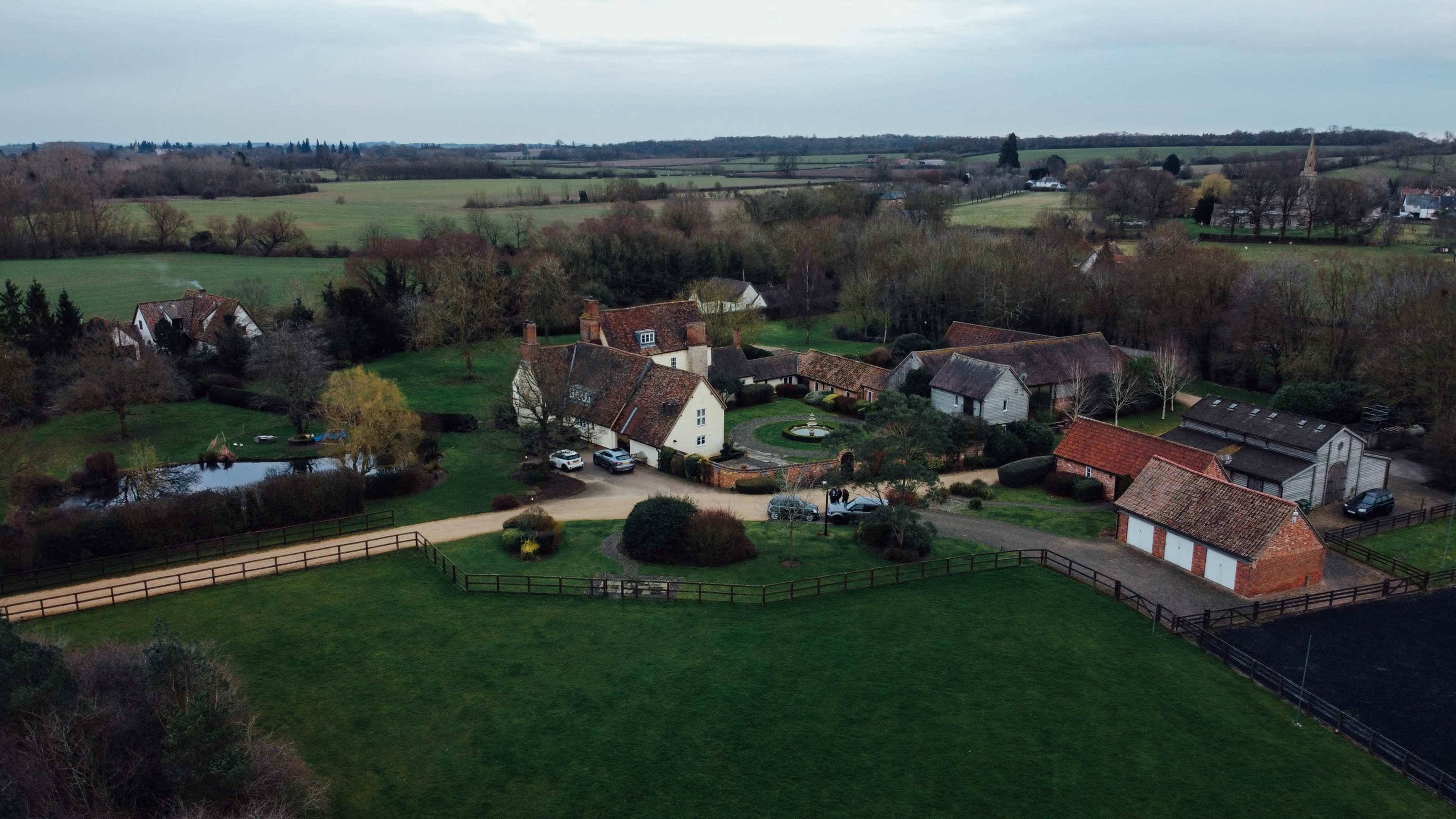 An aerial view shows a cluster of farmhouses and outbuildings surrounded by green fields and trees.