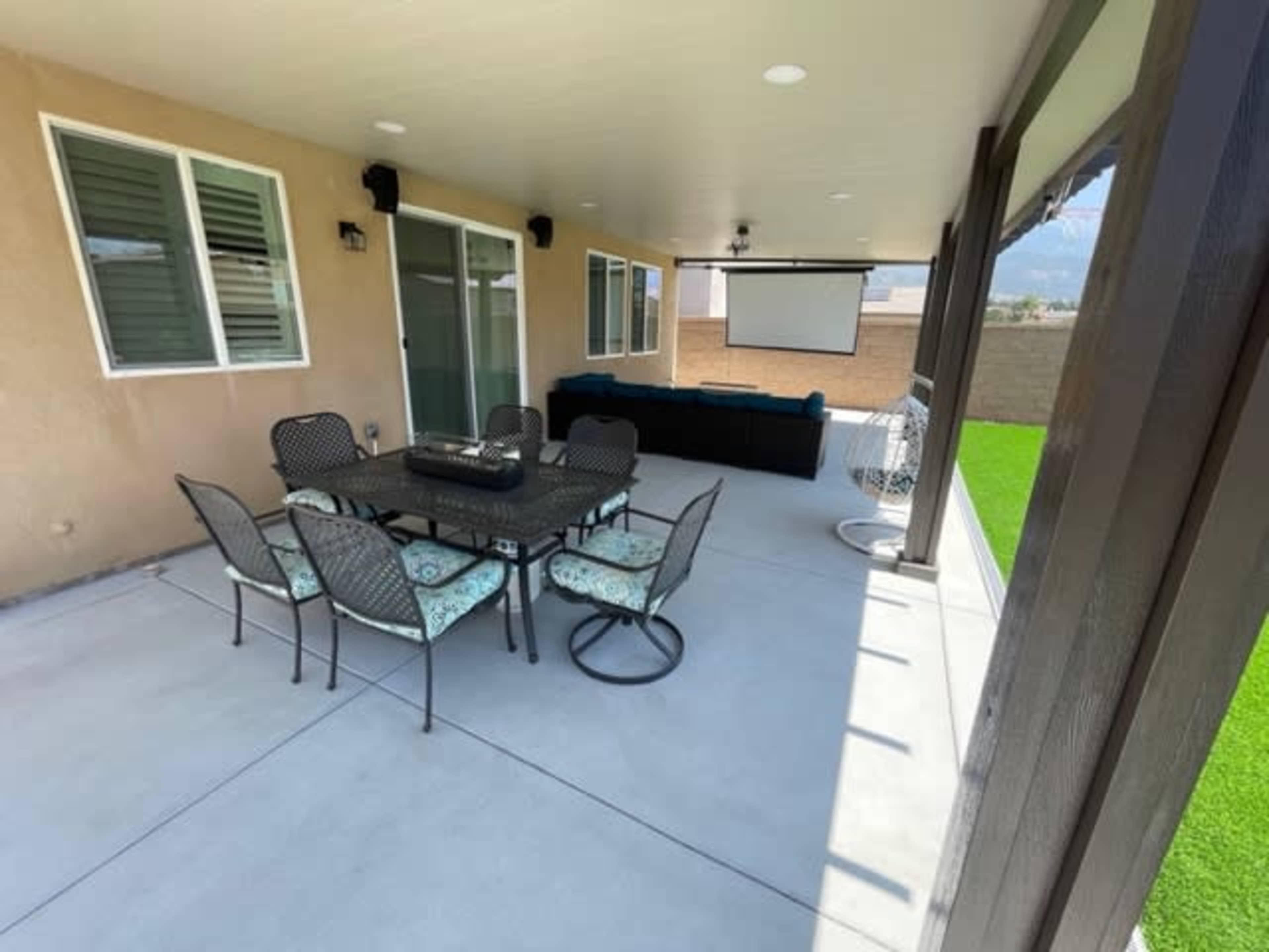 The image shows a covered outdoor patio with a dining table and chairs, a couch, and a projector screen mounted on the wall.