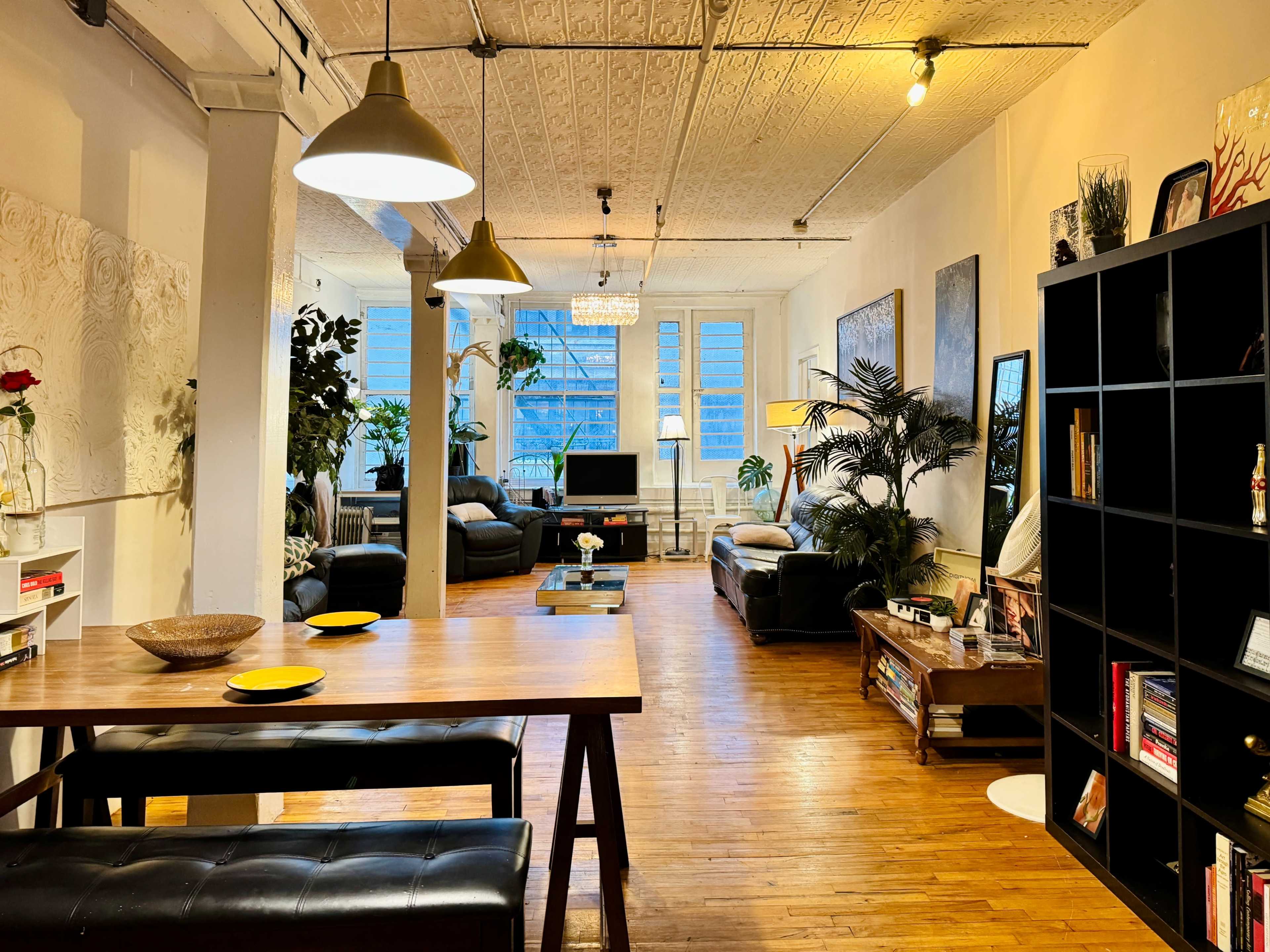 The image shows a spacious living area with hardwood floors, a dining table, leather sofas, decorative plants, and large windows letting in natural light.