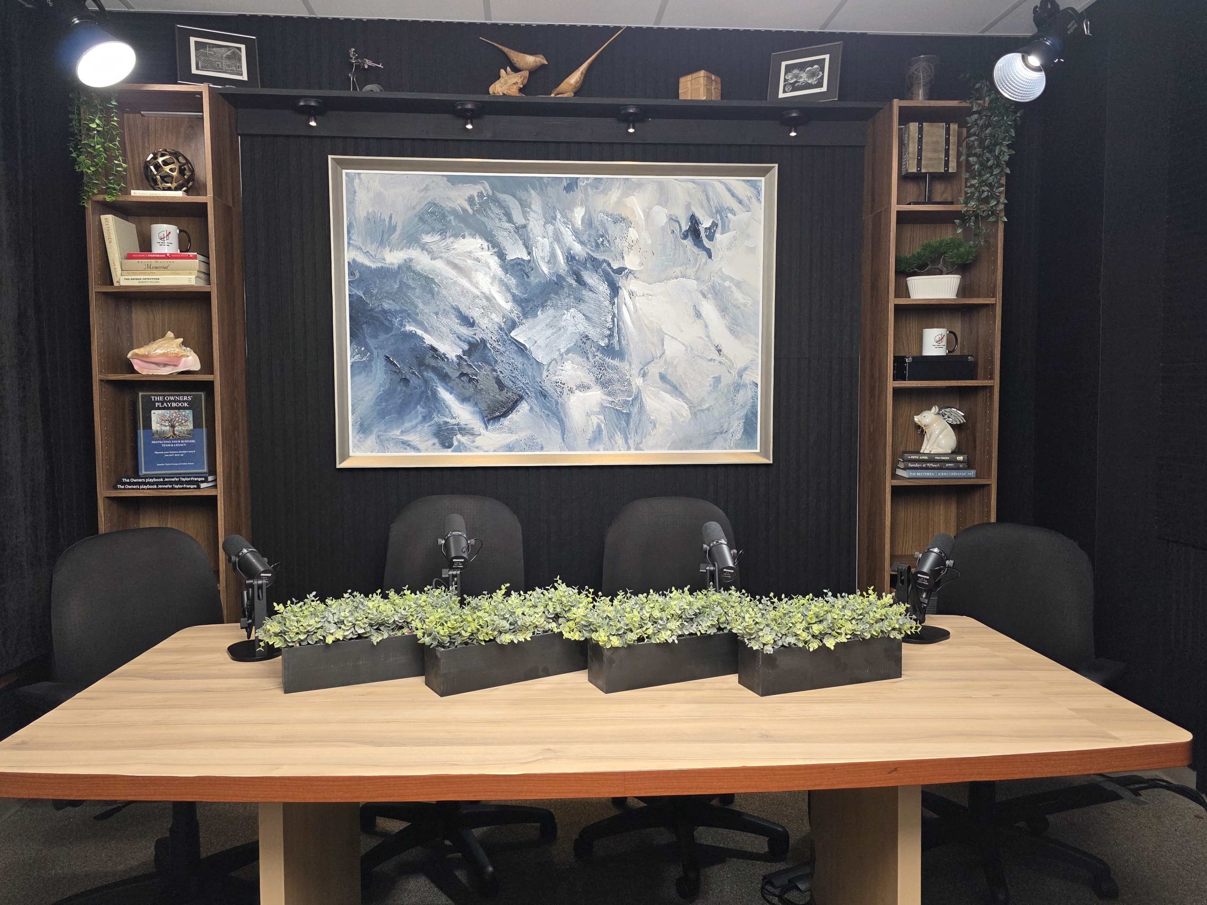 The image shows a modern recording studio with a wooden table, three black chairs, and a backdrop featuring an abstract painting and shelves with decorative items.