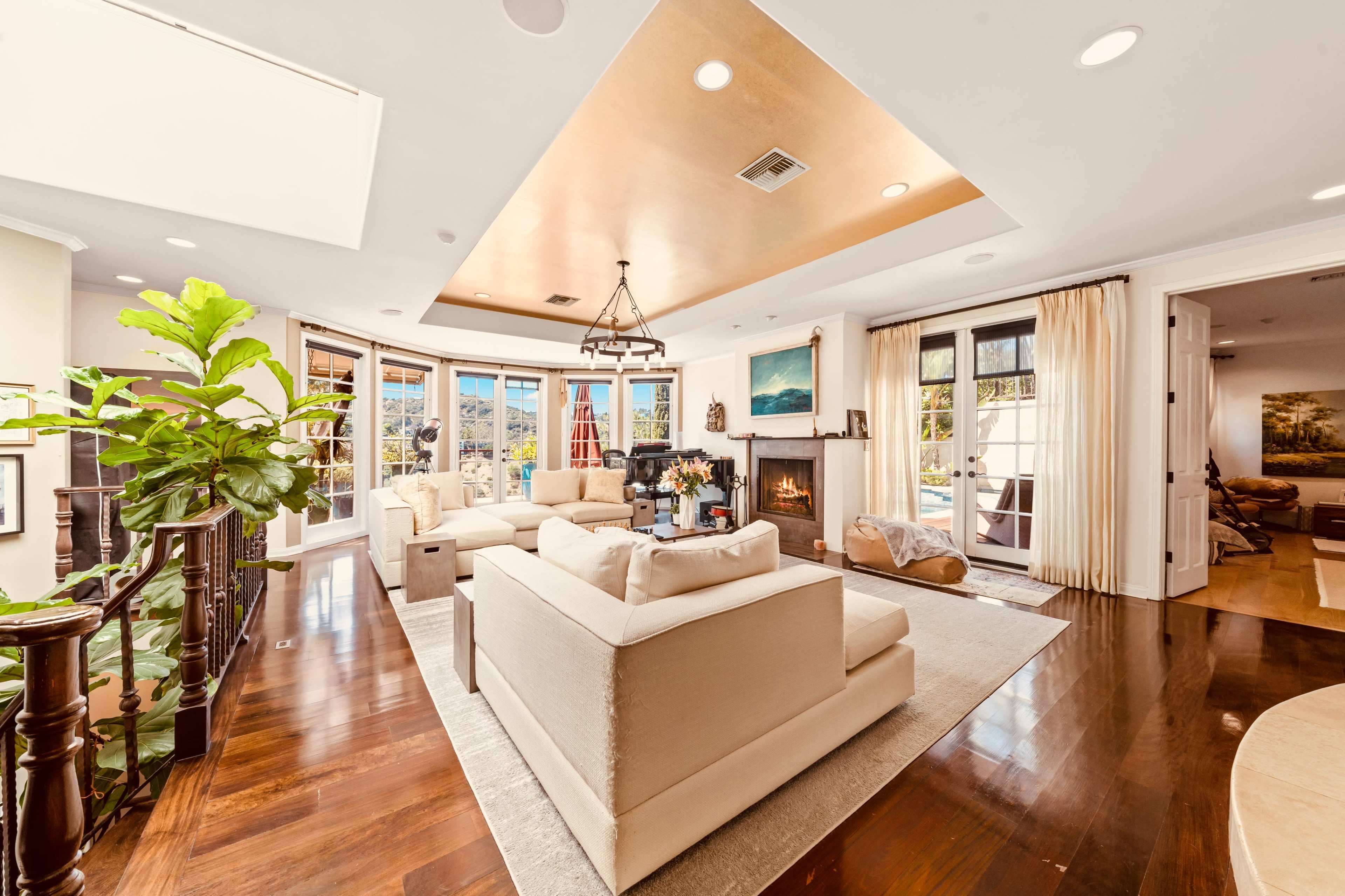 The image shows a spacious living room with a light-colored seating arrangement, a fireplace, large windows, and polished wooden floors.