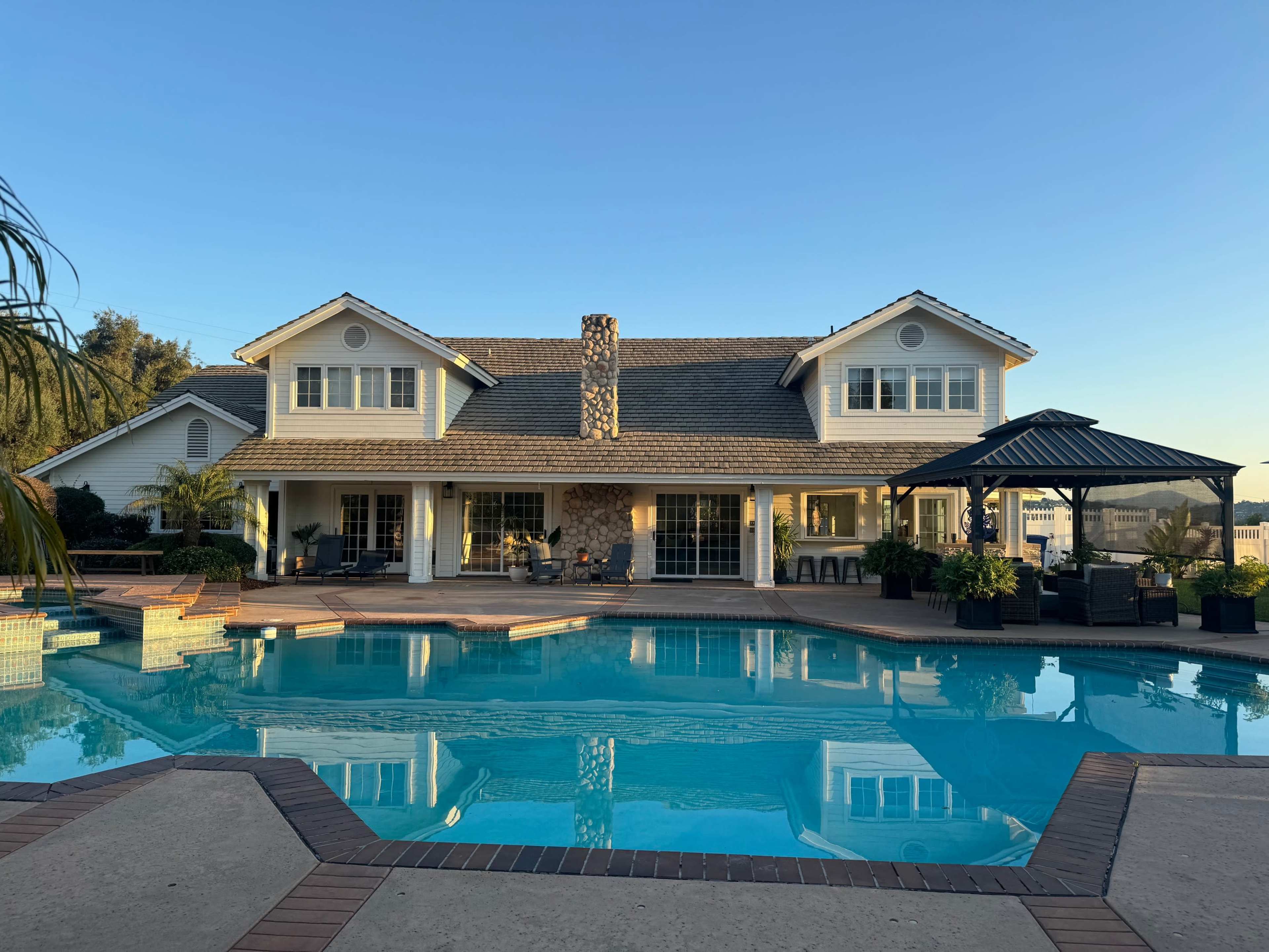 A large, two-story house with a stone chimney and a swimming pool surrounded by a patio and a gazebo.