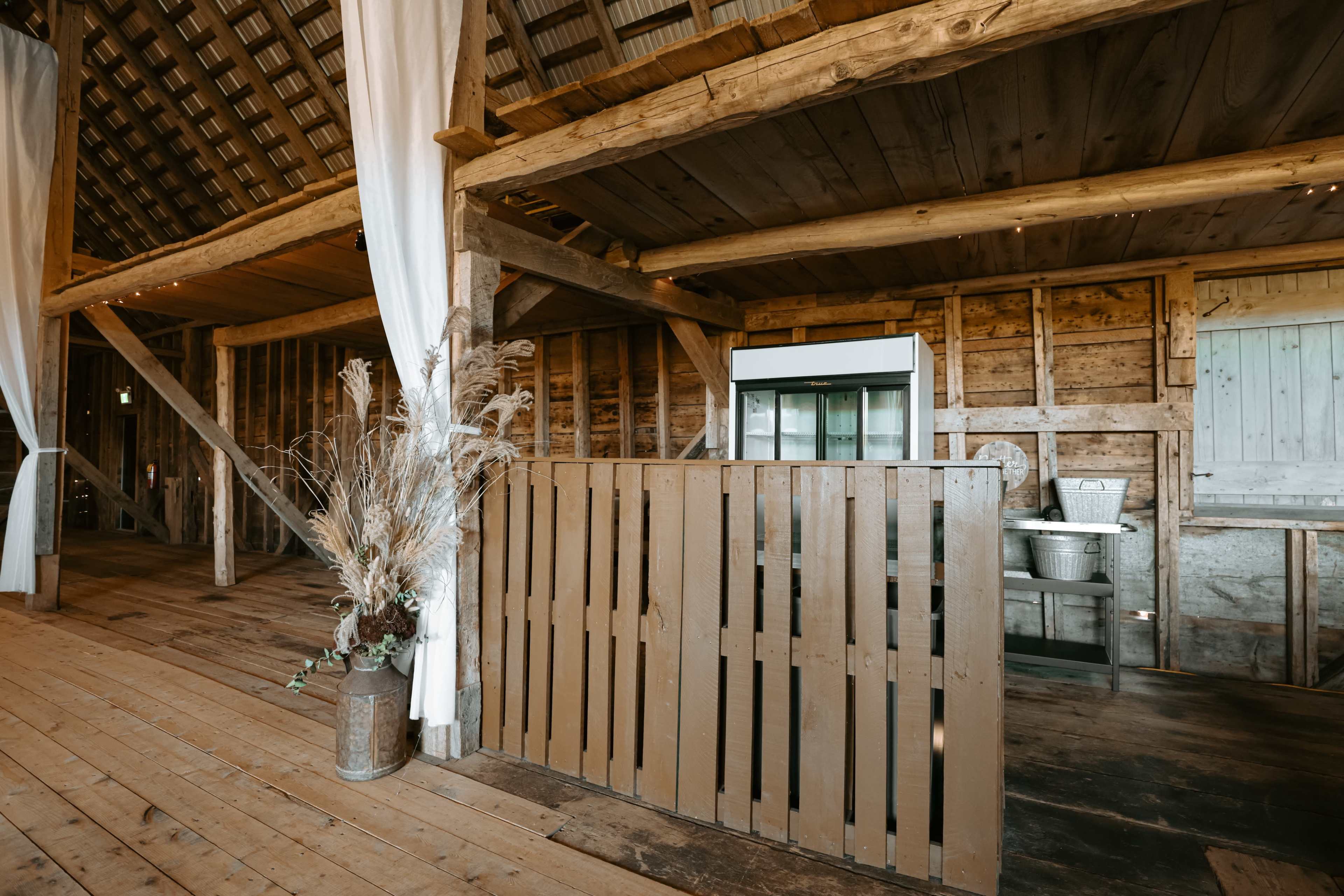 The interior of a rustic barn features wooden walls, a wooden floor, and a partition made of vertical slats, with minimalist décor including dried plants in a vase.