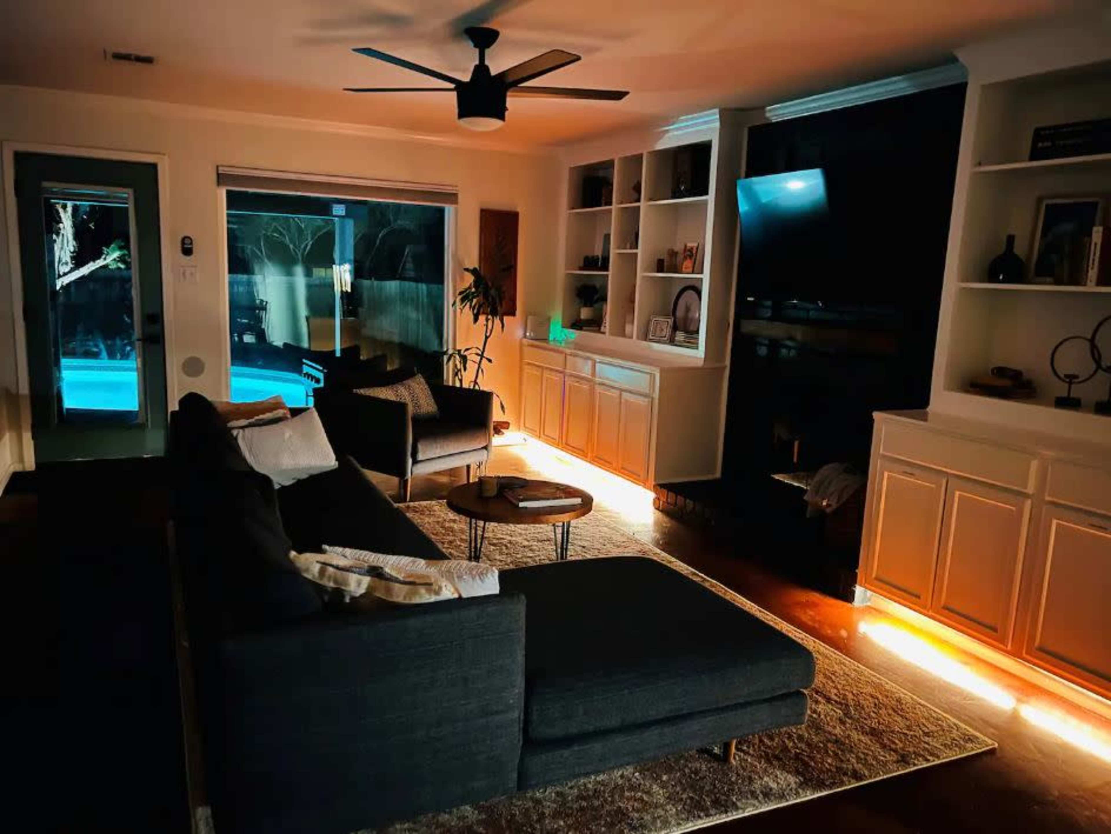 A modern living room is illuminated by soft light, featuring a dark sectional sofa, a round table, and bookshelves, with a view of a pool through large windows.